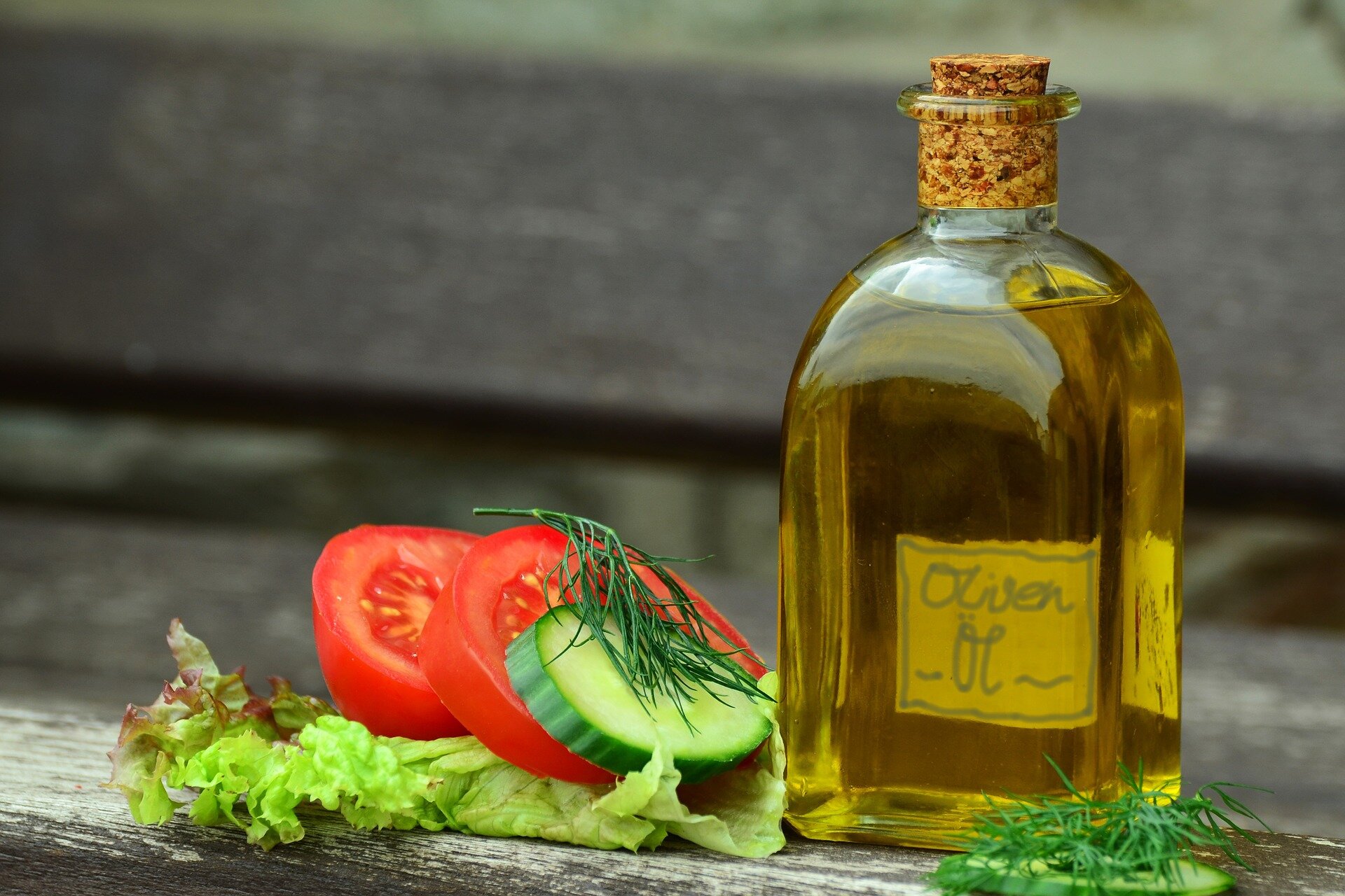 Na zdjęciu jest oliwa w butelce. Obok leżą plasterki pomidora i zielonego ogórka oraz liście zielonej sałaty.