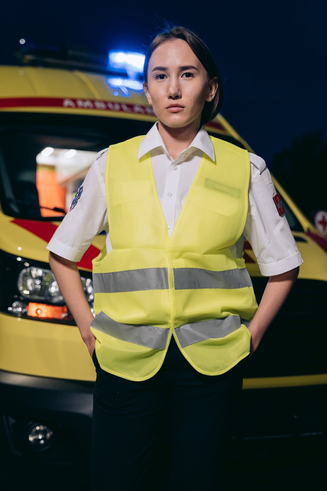 Zdjęcie przedstawia młodą kobietę będącą ratownikiem medycznym. Kobieta jest ubrana w białą koszulę z naszywkami na ramionach oraz w kamizelkę odblaskową. Kobieta stoi na tle ambulansu.