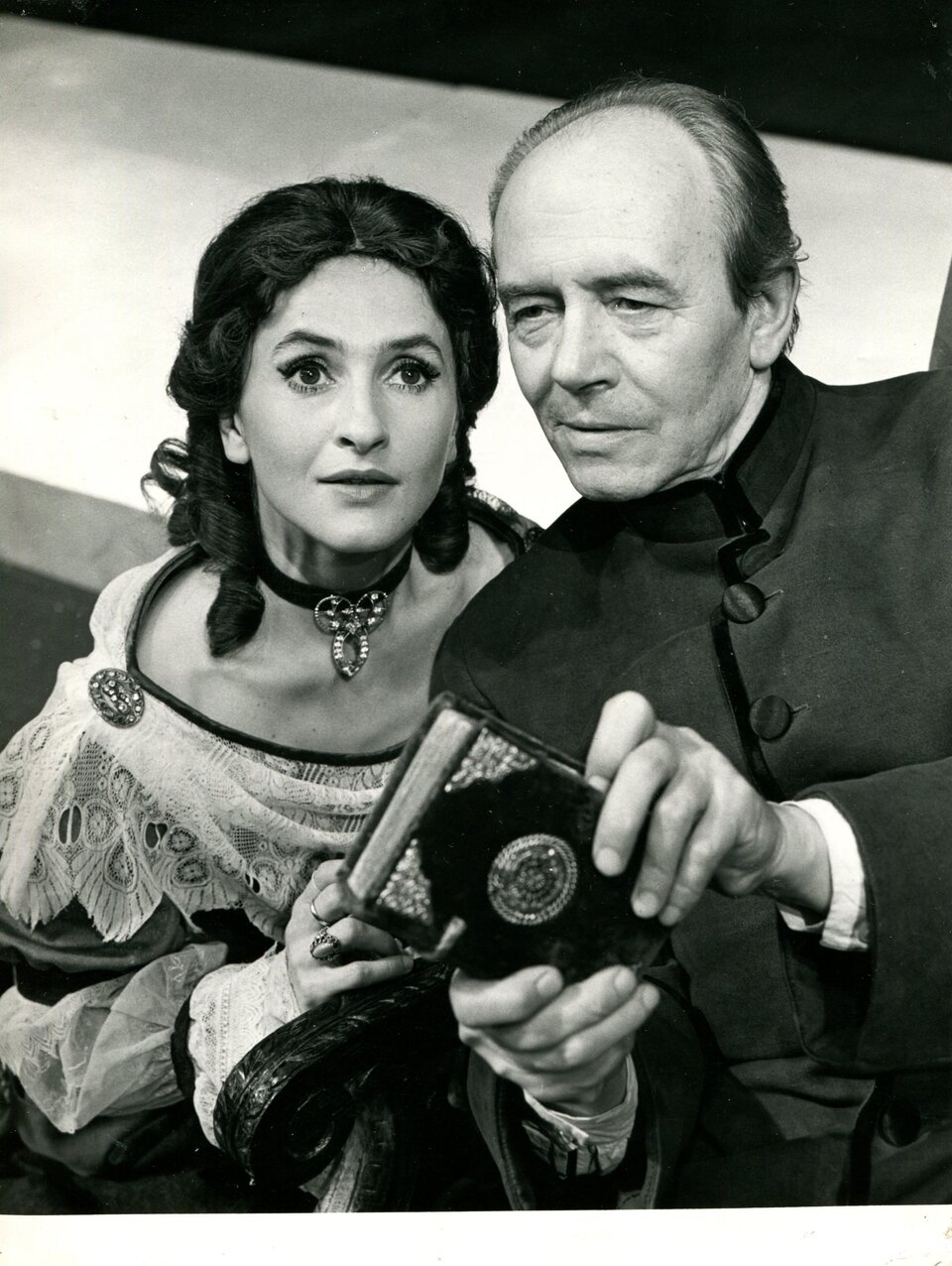 Fotografia przedstawia kobietę i mężczyznę znajdujących się obok siebie. Dojrzała kobieta ukazuje owalną twarz w całości. Postać ma gładkie czoło, ciemne brwi, duże oczy, prosty nos i wydatne usta. Patrzy przed siebie. Kobieta ma ciemne włosy, sięgające ramion. Na szyi ma wąską aksamitkę. Jest ubrana w suknię z dekoltem w kształcie łódki. Mężczyzna jest dojrzały. Ukazuje lewy profil. Postać ma znaczną łysinę. Jego brwi są ciemne, oczy zwężone i otoczone siatką zmarszczek. Mężczyzna ma okazały nos i skrzywione usta. W dłoniach trzyma zamkniętą książkę. Jest ubrany w marynarkę.