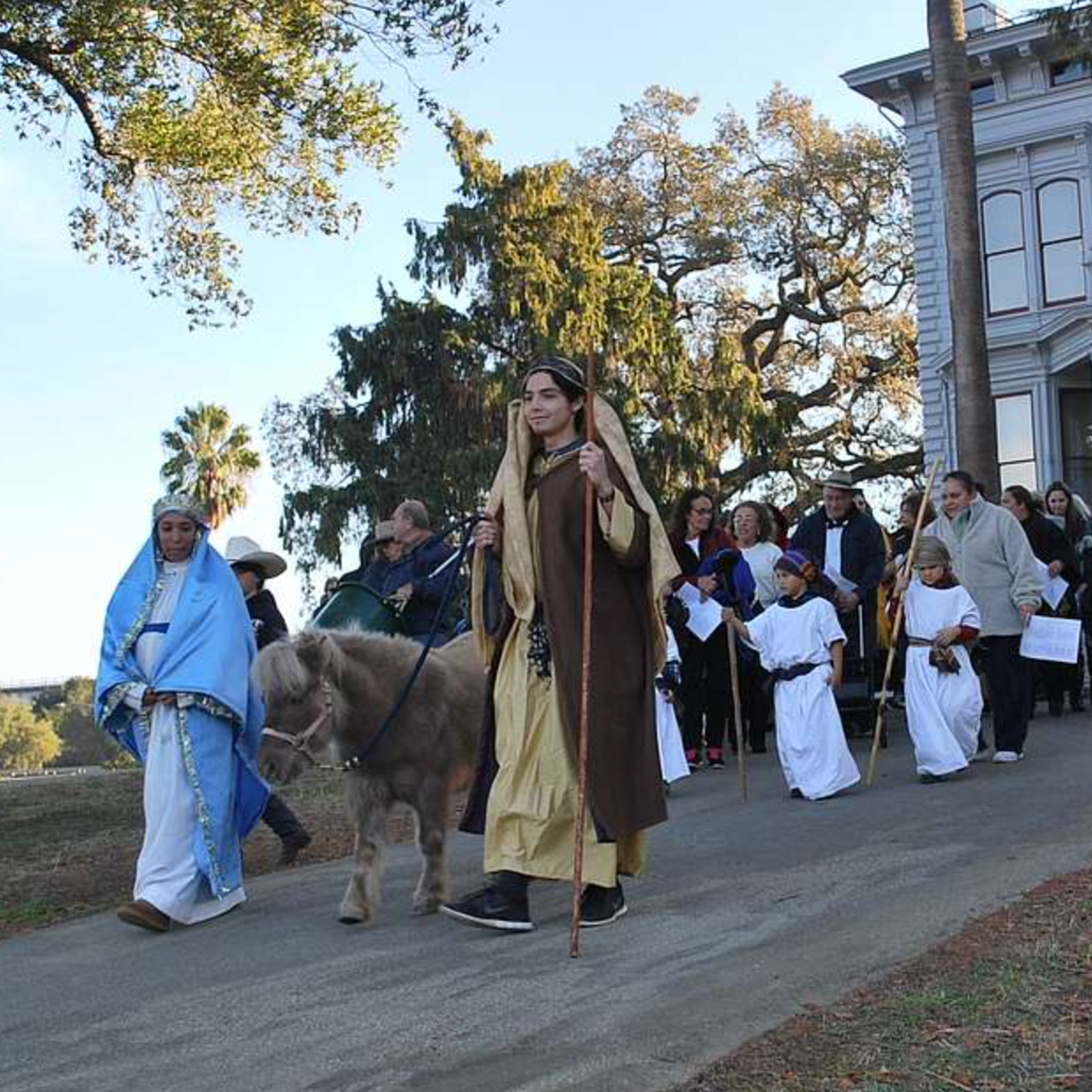 Zdjęcie przedstawia procesję z okazji Bożego Narodzenia. Widoczni są uczestnicy procesji. Na pierwszym planie widoczne są dwie osoby. Ta po prawej ubrana jest w niebieską ozdobną szatę, a ta po lewej w jasnobrązową szatę z kamizelką z kożucha z długim kijem w lewej dłoni. Postać ta prowadzi osiołka. Z tyłu widoczne są osoby dorosłe oraz dzieci ubrane w białe szaty.