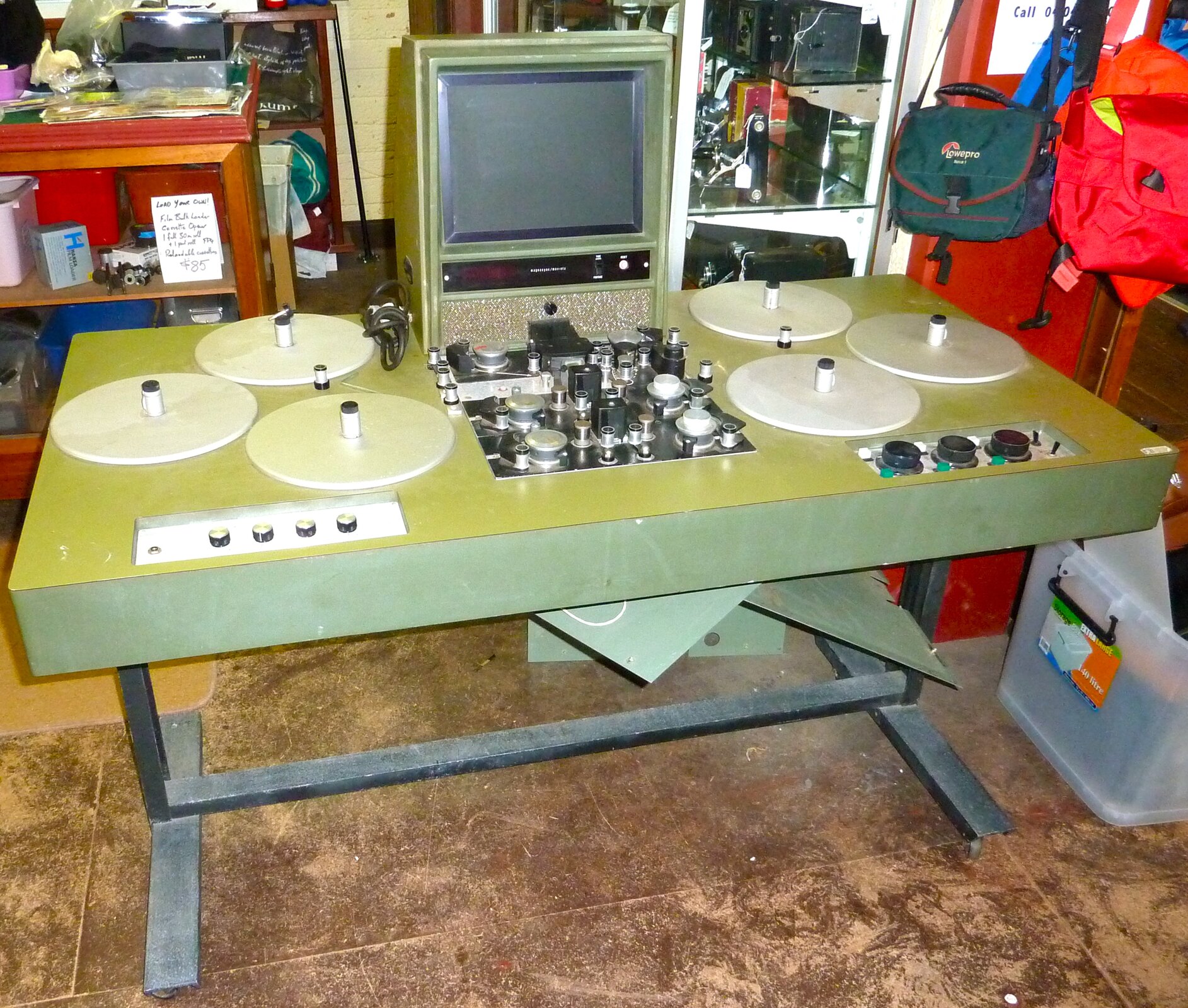 Zdjęcie przedstawia Moviolę, czyli stół do edycji dźwięku na taśmie szesnaście milimetrów. Po lewej i po prawej stronie na stole rozmieszczone są po trzy talerze, z których środka ku górze wystają pręty, na które nakładane są szpule z taśmą. Z lewej i z prawej strony z przodu zamontowane są pokrętła. Na środku znajdują się przyciski, pokrętła i przełączniki, umożliwiające edycję ścieżki znajdującej się na taśmie. Za nimi zamontowany jest monitor.  