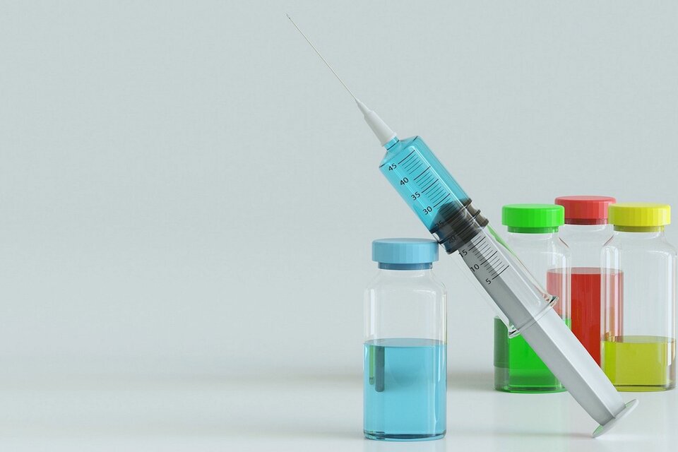 Zdjęcie przedstawia strzykawkę z igłą oraz małe szklane butelki z surowicą. W butelkach znajduje się niebieski, zielony, jasnozielony i czerwony płyn. 