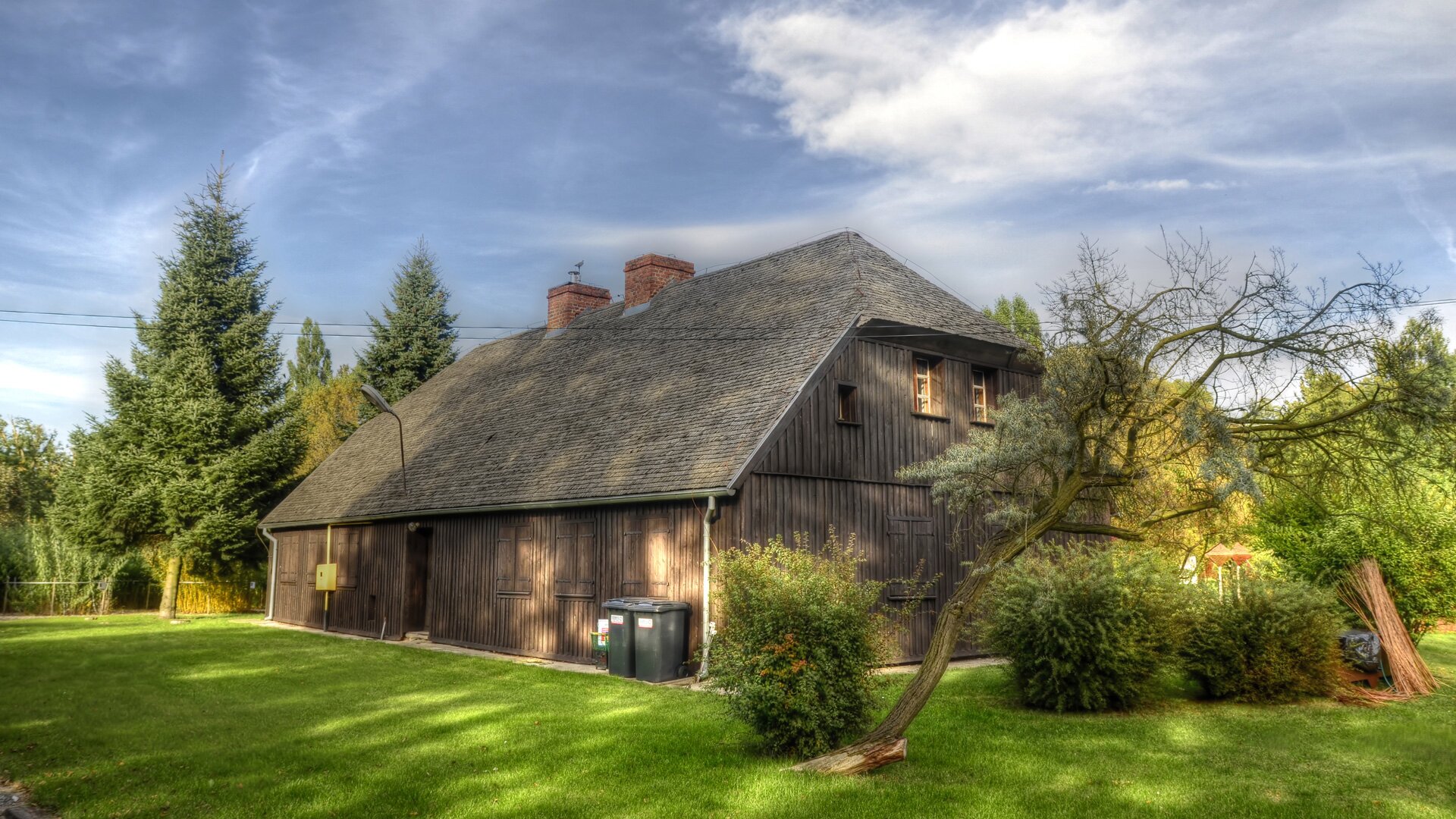 Zdjęcie przedstawia drewniany prostokątny dom przykryty drewnianym dachem. Otoczony jest drzewami.