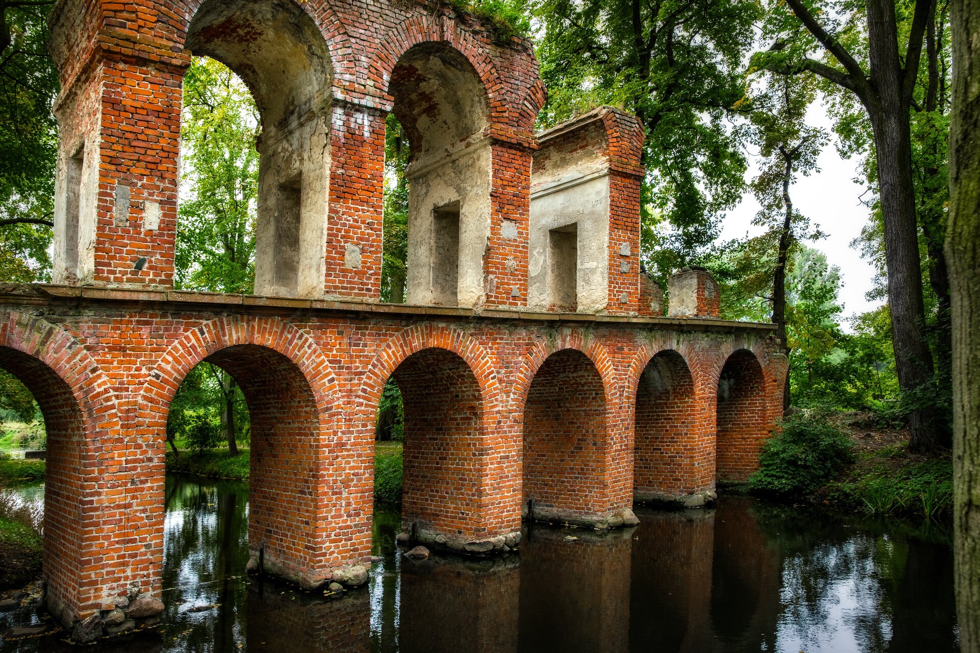 Ilustracja przedstawia Akwedukt na rzece Skierniewce w polskiej wsi Arkadia. Ukazuje ceglany most łączący brzegi rzeczki tuż przy ujściu do wielkiego stawu. Przęsła wykonane są z łuków i ułożone są jedno nad drugim. W tle rosną drzewa, przez które prześwituje niebo. Obiekt wzorowany jest na starożytnych akweduktach.