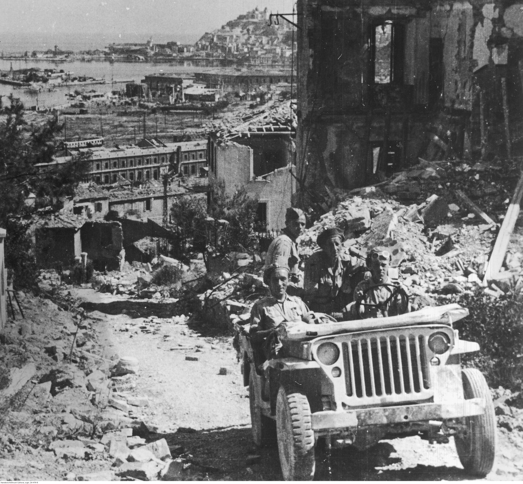 Zdjęcie przedstawia czterech polskich żołnierzy jadących Jeepem po ulicach zniszczonej Ankony. Wkoło nich widać liczne zbombardowane budynki. W tle widać port.