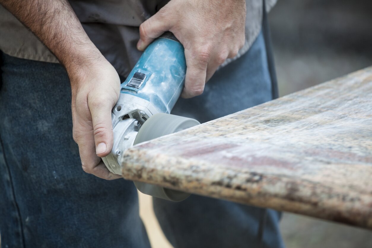 Zdjęcie przedstawia granitową płytę o nieregularnej strukturze. Kamieniarz specjalnie przeznaczoną do tego szlifierką poleruje brzeg płyty.