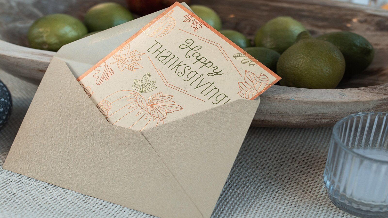Zdjęcie przedstawia szarą, otwartą kopertę, z której wystaje kartka okolicznościowa. Na kartce widoczny jest napis w języku angielskim: HAPPY THANKSGIVING oraz rysunki dyni i liści. Kartka oparta jest o płaską miskę wypełnioną zielonymi limonkami. Po prawej stronie widoczny jest szklany pojemnik z białym płynem na dnie. 