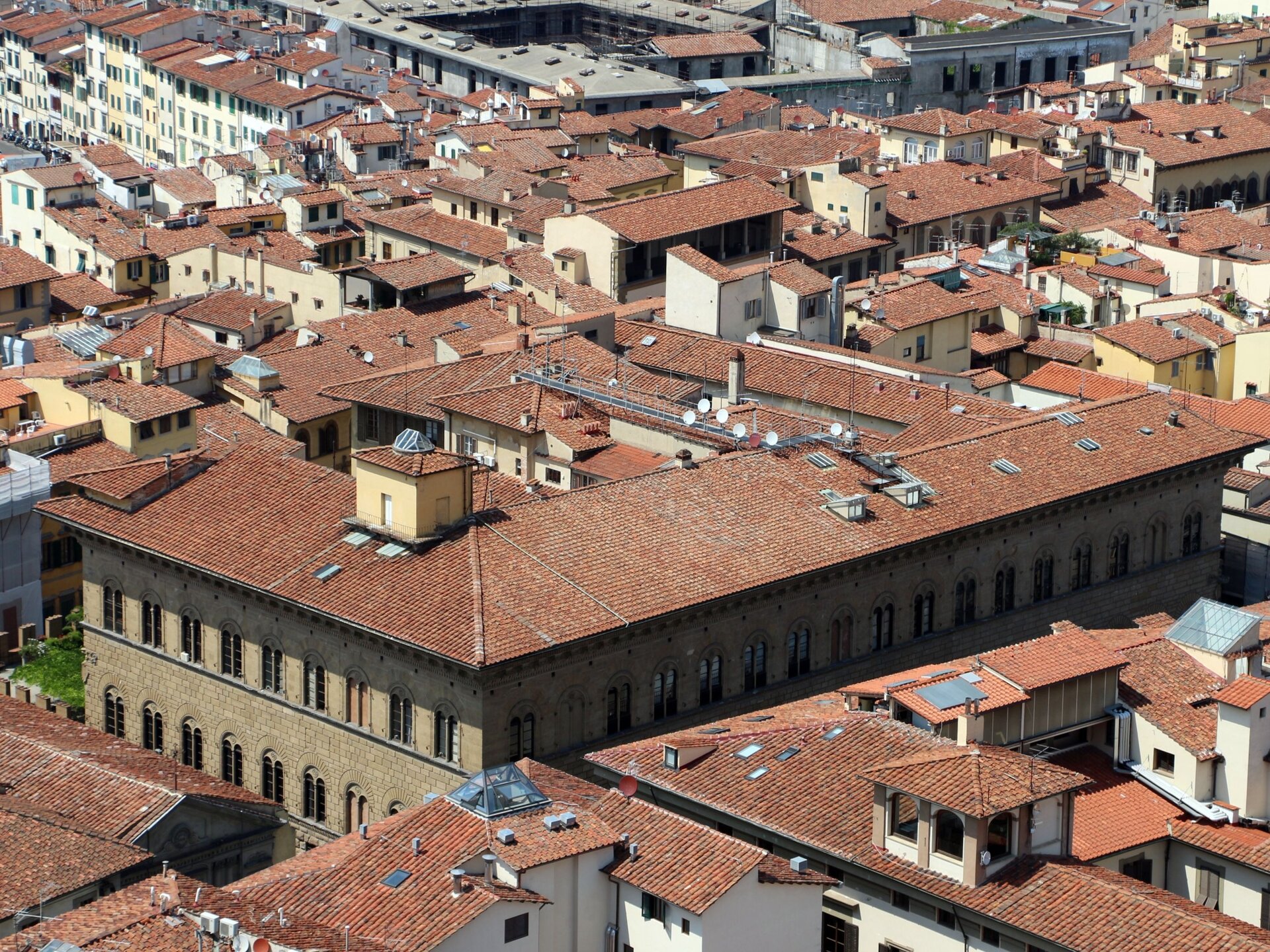 Fotografia barwna przedstawiająca Palazzo Medici we Florencji. Dach pałacu, jak i innych domów jest niemal płaski i pokryty pomarańczowymi dachówkami. Okna są zbliżone kształtem do prostokątów z delikatnym łukiem ponad nimi.