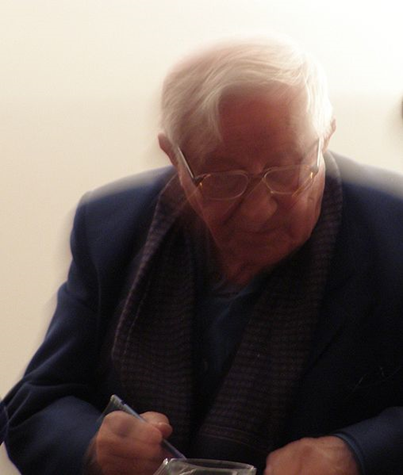 Zdjęcie przedstawia starszego mężczyznę. Ma siwe, zaczesane na bok włosy, zmarszczki na twarzy oraz założone okulary. Ubrany jest w ciemną koszulę i marynarkę. W prawej ręce trzyma długopis. Zdjęcie jest lekko rozmazane.