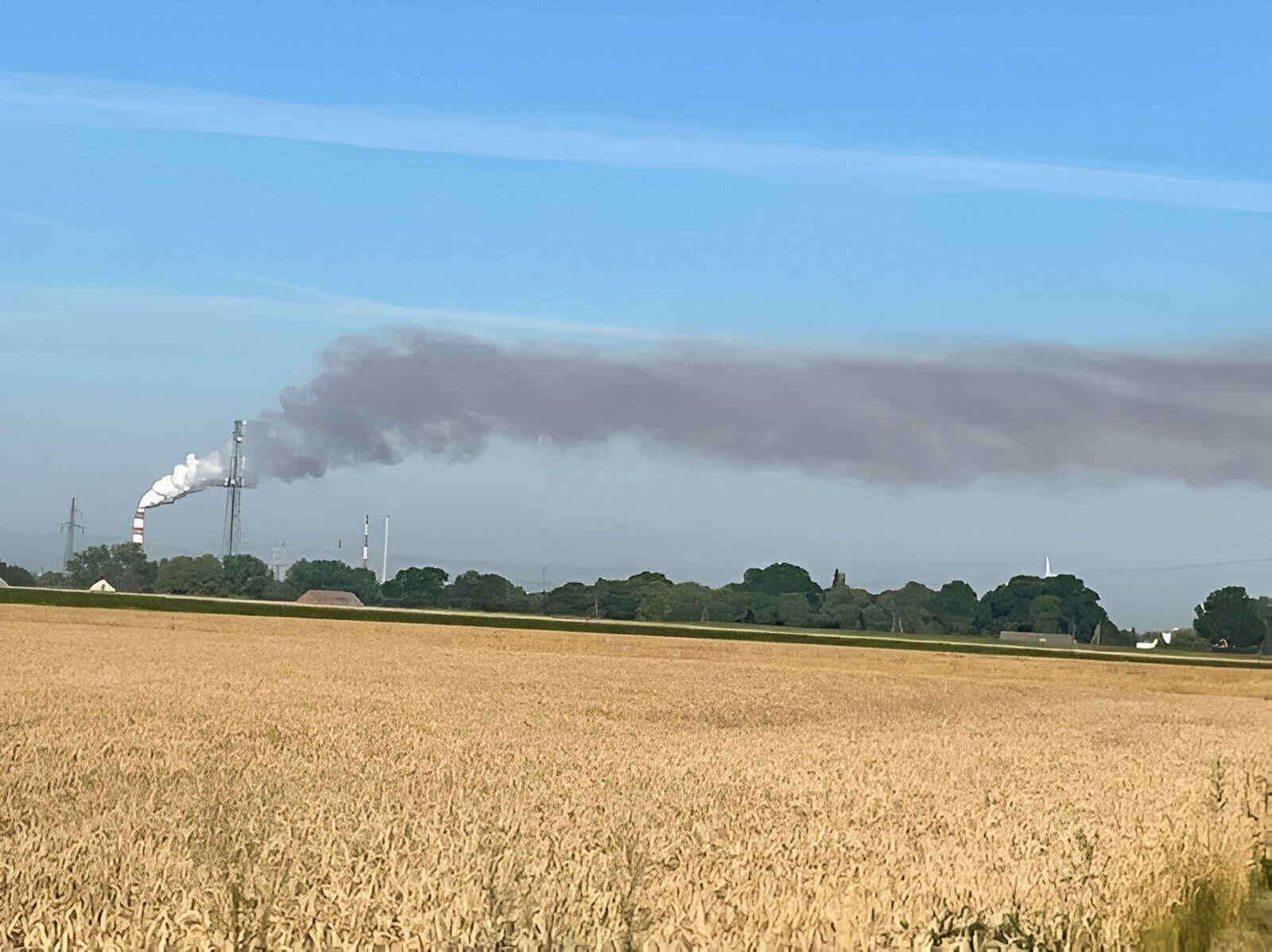 Fotografia wykonana na terenie podmiejskim. Na pierwszym planie pole zboża, w oddali linia drzew, a dalej, po lewej stronie, wysoki komin kombinatu, z którego wydobywa się bardzo duża ilość gęstego dymu. Na początku jest biały, następnie smuga rozszerza się w prawą stronę i przyjmuje szary kolor. Ciągnie się na setki metrów.