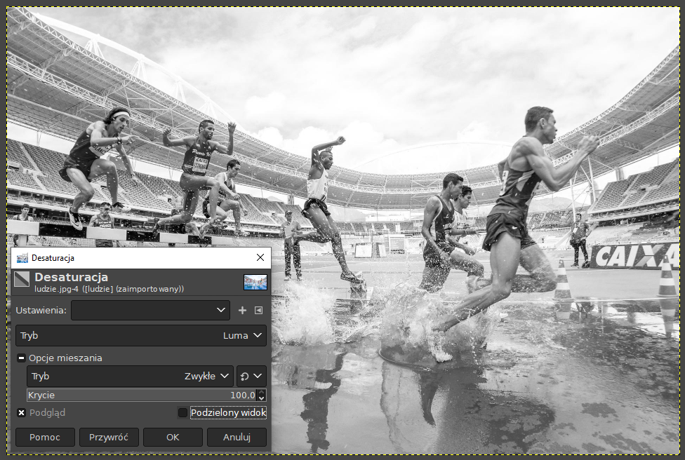 Ilustracja przedstawia okno programu. W obszarze roboczym jest zdjęcie biegnących zawodników na stadionie. Zdjęcie jest utrzymane w odcieniach szarości. Na tle zdjęcia jest okno o nazwie: Desaturacja. W oknie są odpowiednie parametry, między innymi Tryb - Luma, w Opcjach mieszania Tryb - Zwykłe, Krycie 100,0. Zaznaczono: Podgląd.  