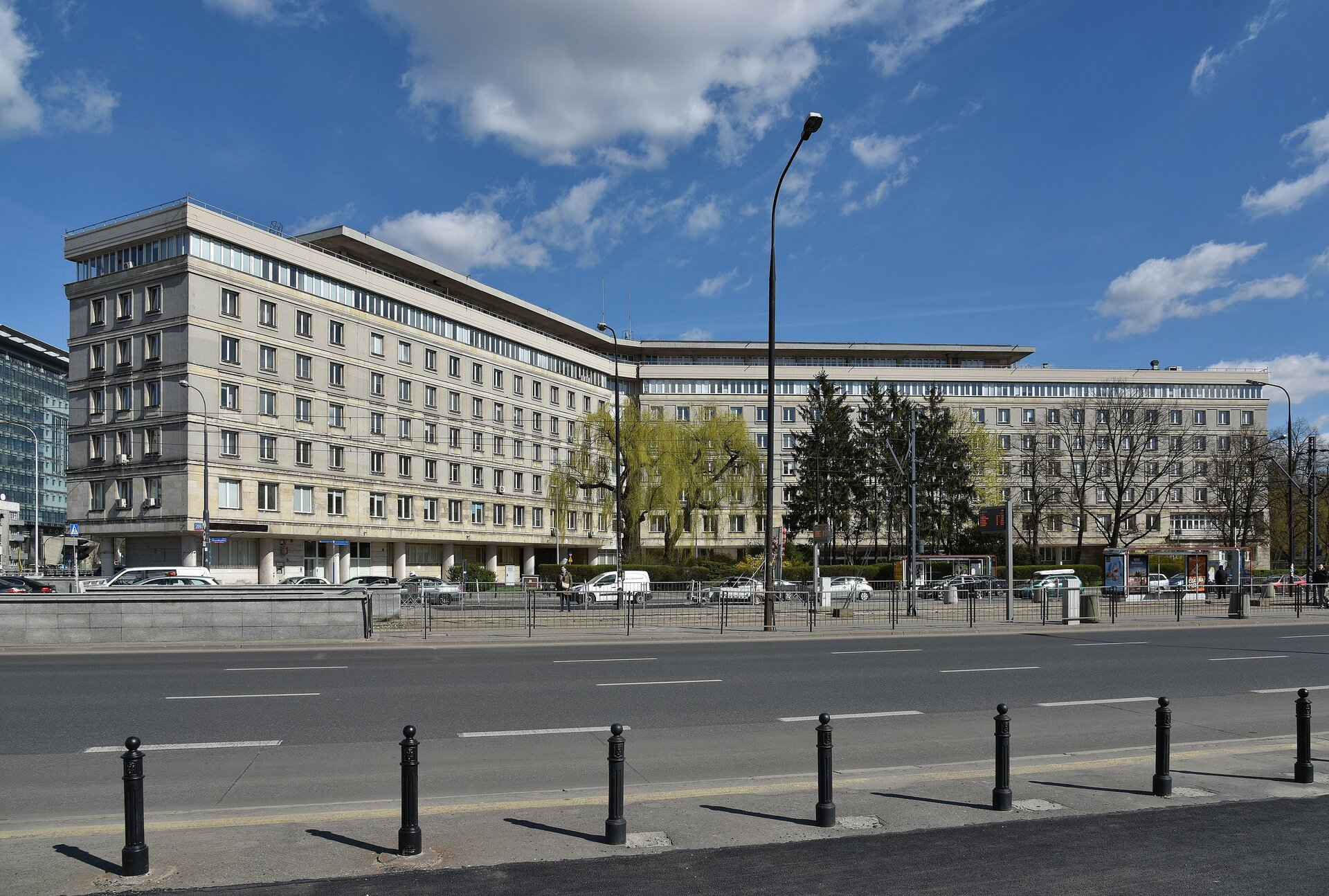 Zdjęcie przedstawia siedzibę GUS w Warszawie. To wysoki, kilkupiętrowy, dwuskrzydłowy budynek stojący przy szerokiej ulicy.