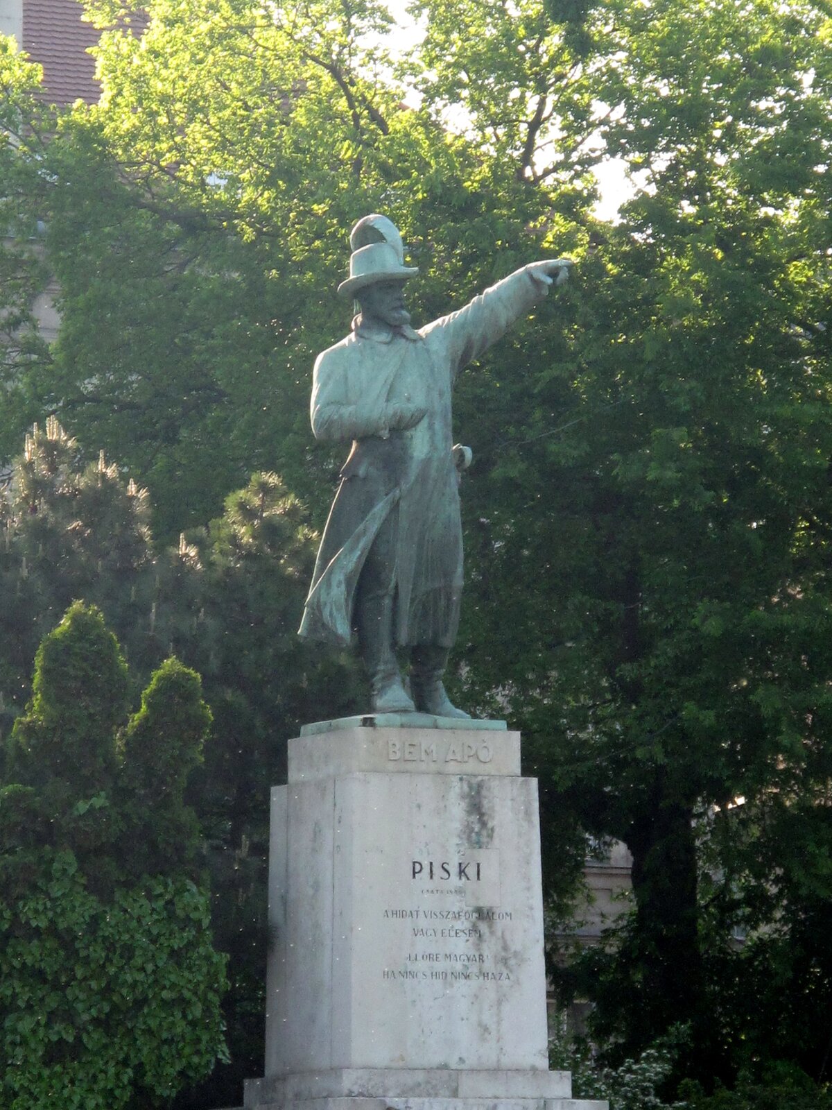 Na zdjęciu pomnik przedstawiający stojącego mężczyznę na postumencie. Ubrany jest w płaszcz oraz kapelusz. Ma uniesioną lewą rękę, którą coś wskazuje. W tle rosną drzewa.