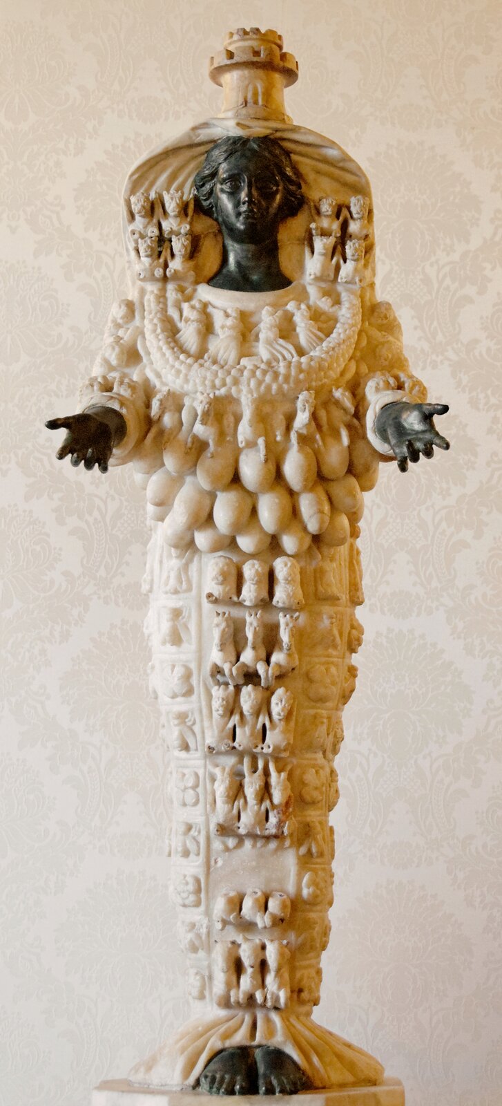 Ilustracja przedstawia rzeźbę Artemidy z Efezu. Głowa, ręce oraz stopy są w kolorze czarnym. Ubrana jest jakby w białą suknię, na której znajdują się małe rzeźby zwierząt. Kobieta jest przestawiona z wieloma piersiami, trzy rzędy piersi umieszczone zostały w dolnej części korpusu.