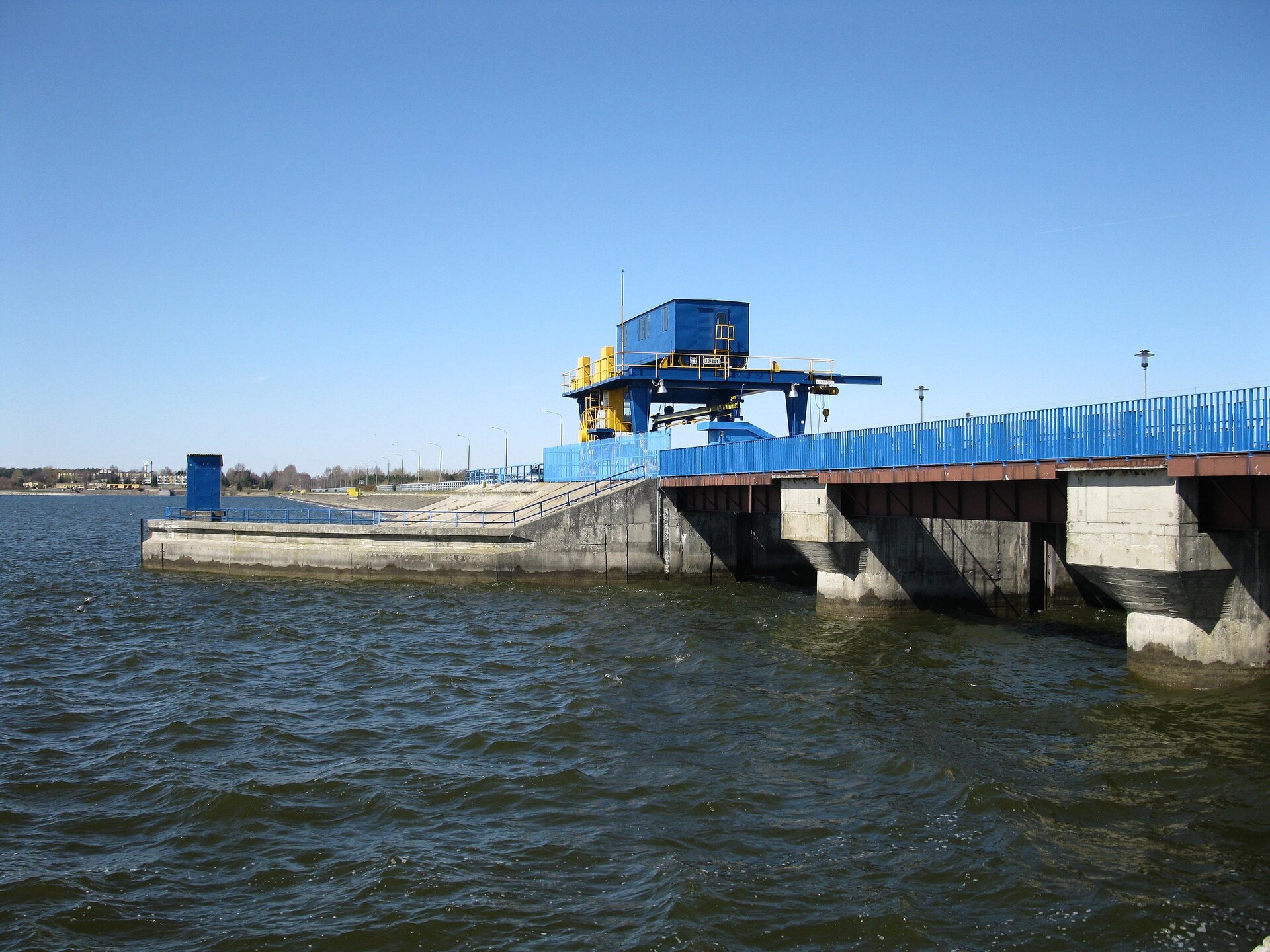 Zdjęcie przedstawia betonową konstrukcję tamy na rzece. U góry biegnie przejście dla pieszych z barierkami po bokach i lampami. Pośrodku konstrukcji kontener na podwyższeniu otoczony barierkami.