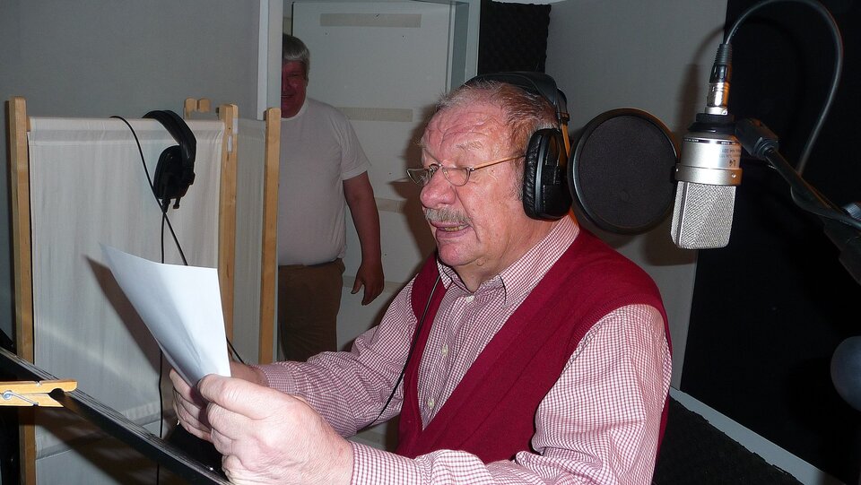 Zdjęcie przedstawia starszego mężczyznę znajdującego się w studiu. Mężczyzna ma założone na uszy słuchawki, siedzi przy mikrofonie, trzyma w dłoniach kartkę papieru.