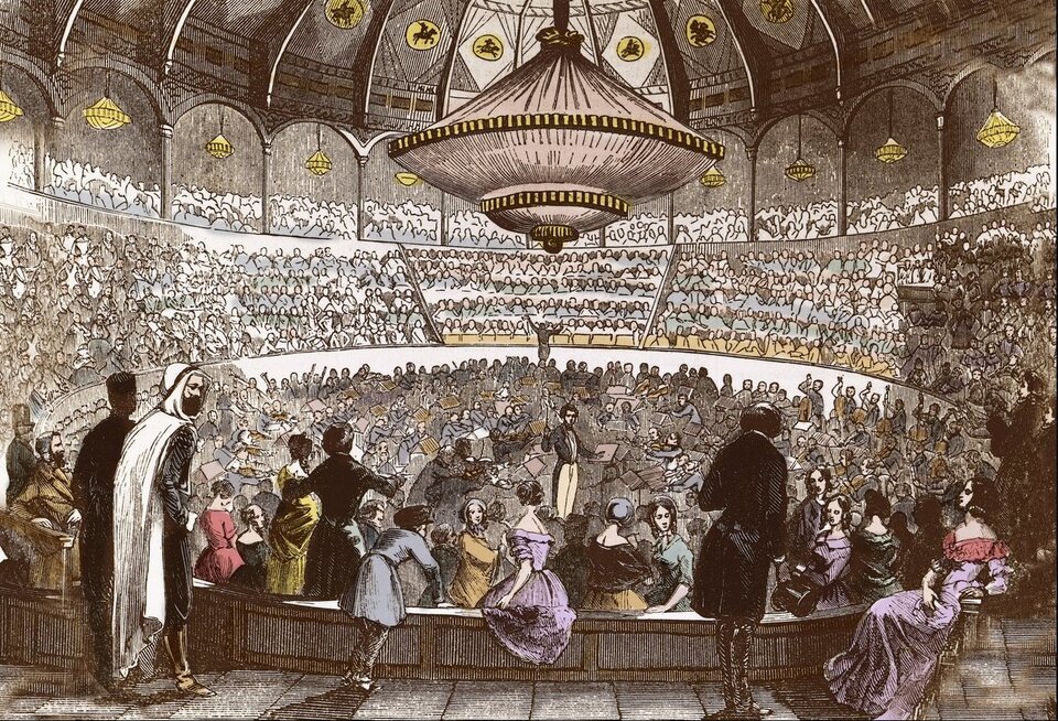 Ilustracja przedstawia tłum ludzi na sali koncertowej. Ludzie siedzą zarówno na balkonach jak i w części dolnej sali. Sufit jest bardzo wystawny i bogato zdobiony.