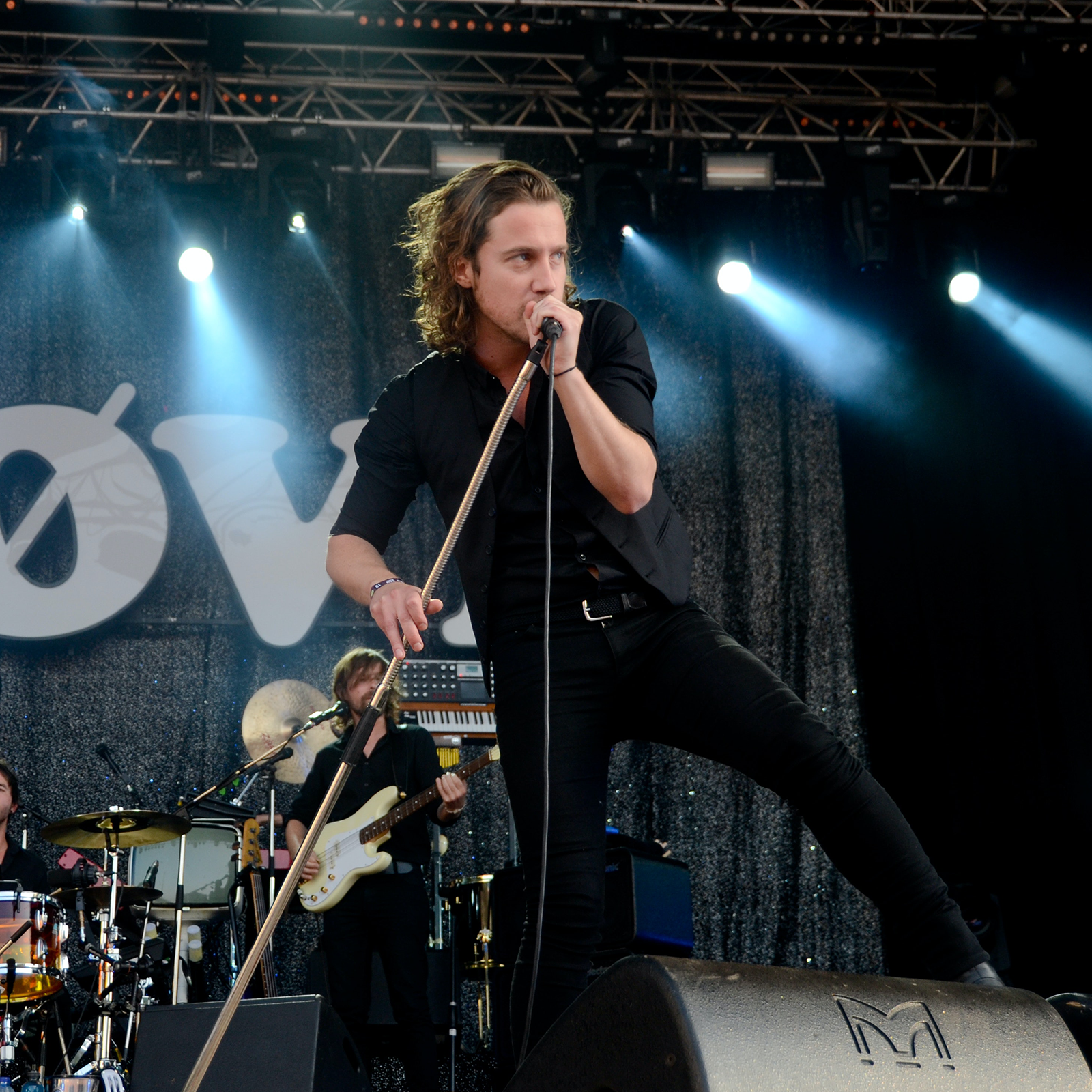 Zdjęcie przedstawia młodego mężczyznę o długich włosach, ubranego w czarną koszulę i czarne spodnie, stojącego na scenie i trzymającego w rękach mikrofon, który przybliżył do ust. W tle widoczny jest mężczyzna grający na gitarze. Scenę oświetlają reflektory przymocowane do metalowej konstrukcji. 