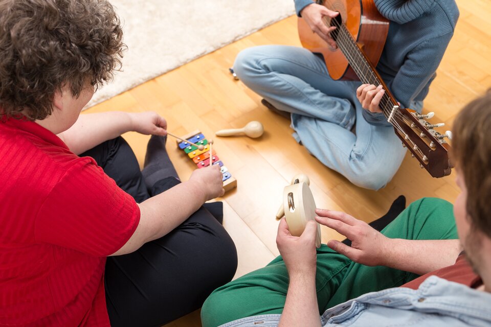 Zdjęcie zatytułowane Muzykoterapia aktywna. Na zdjęciu trzy osoby - jedna gra na gitarze, druga na dzwonkach, trzecia na tamburynie. Osoby siedzą po turecku, w kręgu, odwrócone są do siebie twarzami.