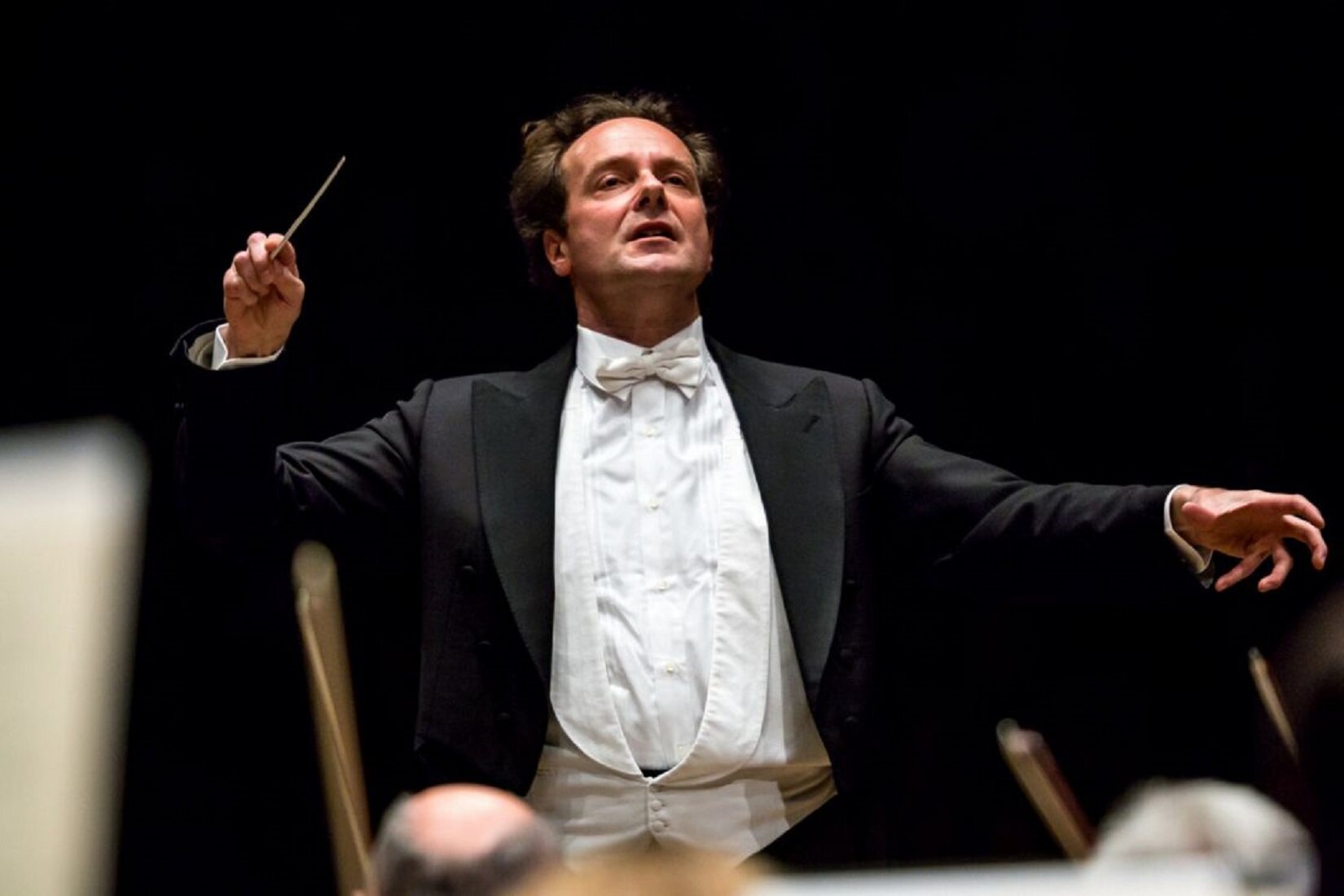 Zdjęcie przedstawia mężczyznę - dyrygenta przy pracy. Mężczyzna w prawej, uniesionej dłoni trzyma batutę, a lewą rękę ma wyciągniętą do boku. Głowę unosi do góry, a jego wzrok utkwiony jest w dali. Mężczyzna ubrany jest w białą koszulę, białą kamizelkę, białą muszkę i czarną marynarkę. Przed nim siedzi orkiestra.