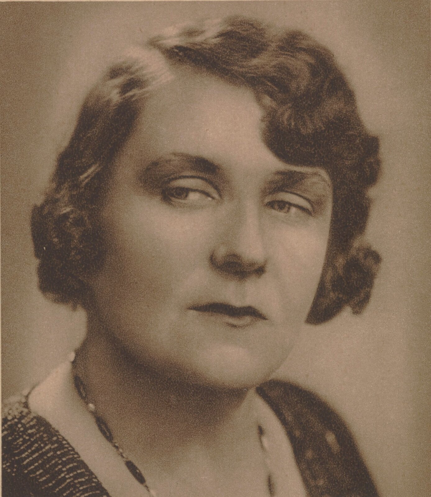 Zdjęcie przedstawia portret dojrzałej kobiety. Ma ona niedługie, pofalowane włosy. Są one starannie ułożone wokół twarzy. Kobieta ma okrągłą twarz. Jej oczy są duże, okalają je od góry wyraziste brwi. Kobieta spogląda w swoją prawą stronę. Ma poważny wyraz twarzy. Na jej szyi wisi delikatny naszyjnik.