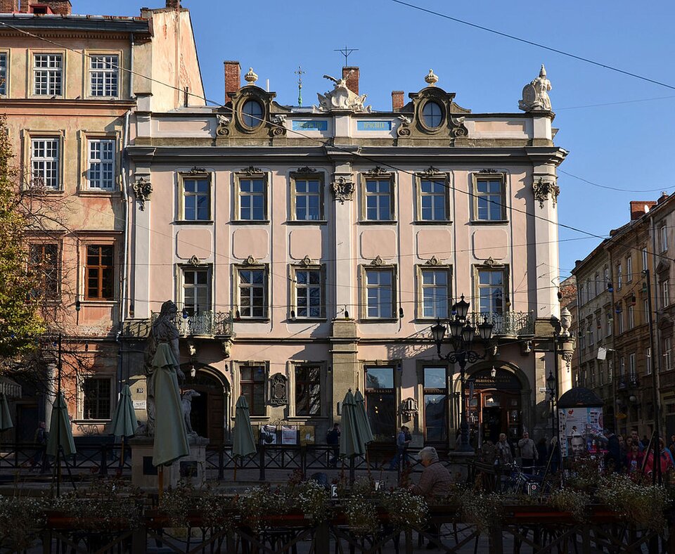 Na fotografii znajduje się Pałac Lubomirskich we Lwowie. To niewielki trzykondygnacyjny budynek o jasnej elewacji z delikatnymi zdobieniami. Budynek posiada duże okna zajmujące większość elewacji oraz rzeźby umieszczone na dachu. Pomiędzy otworami okiennymi znajdują się pilastry.