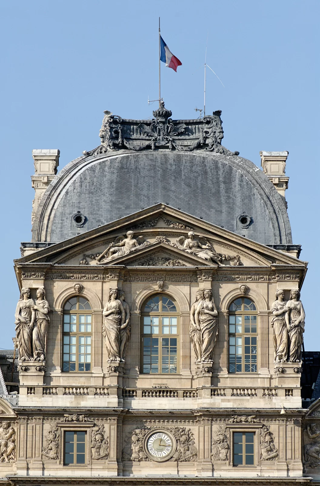 Zdjęcie przedstawia fragment jednej ze ścian budowli. Jest ona bogato zdobiona kamiennymi rzeźbami o różnych kształtach i wzorach. W górnej części budynku znajdują się trzy, łukowo zakończone okna. Pomiędzy nimi znajdują się po dwie rzeźby stojących kobiet w długich szatach. Rzeźby pełnią funkcję kolumn. Na swoich głowach trzymają wyższą część budynku. Postacie ustawione skrajnie po lewej oraz prawej stronie stoją obok siebie. Jedna kładzie drugiej ręce na ramionach. Postacie patrzą na siebie nawzajem. Postacie ustawione bliżej środka znajdują się jedna za drugą. Jedna trzyma drugą za rękę.
