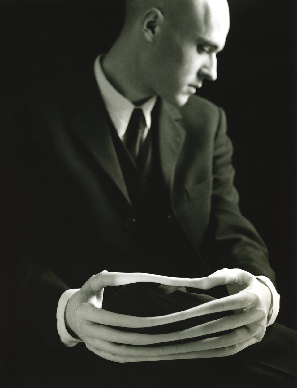 Czarno‑biała fotografia przedstawiająca półpostaciowy portret młodego mężczyzny. Został on ukazany frontem do widza. Ma on pochyloną głowę na bok w lewo i w dół, tak że widać tylko prawą stronę twarzy: prawe ucho, prawe oko, prawą brew, prawą część nosa i ust. Ma on wysokie, jasne czoło i głowę niemal pozbawioną włosów. Mężczyzna jest ubrany w garnitur, białą koszulę, a pod szyją ma zawiązany krawat. Dłonie są zwrócone zewnętrzną stroną do widza, na nich białe rękawiczki mocno przylegające do ciała, które wydłużyły palce tak, że palce jednej ręki stykają się z palcami drugiej. Na twarzy mężczyzny maluje się powaga i skupienie. Tło jest czarne.