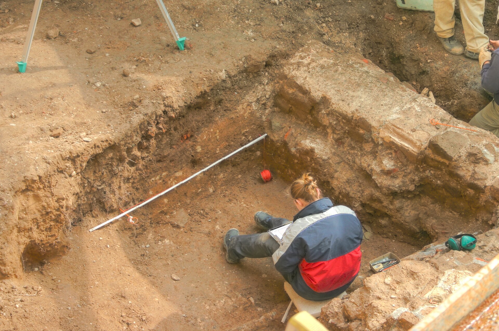 Zdjęcie przedstawia kobietę siedzącą w kwadratowym dole, czytającą coś z kartek trzymanych na kolanach.
