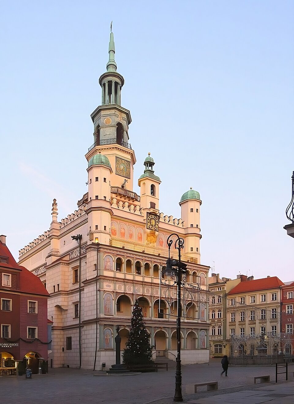 Zdjęcie przedstawia widok współczesny na budynek poznańskiego ratusza, jego fasadę i ścianę boczną. Parter i dwie pierwsze kondygnacje oddzielone gzymsami na fasadzie posiadają arkady o pełnych łukach otworów. Na fryzach przedstawienie postaci męskich w strojach rycerskich i szlacheckich. W górnej części kondygnacji zdwojenie wieżyczek w narożnikach fasady, attyka na całej szerokości fasady budynku , pośrodku wyrastająca ponad szczyt budynku wieżyczka z zegarem tarczowym. Wieża dzwonna z zegarem wieńczy szczyt budynku. Wszystkie wieże pokryte są hełmami od góry. Boczna ściana budynku posiada wykusz i okna prostokątne, ciągnące się pod linią attyki ratusza. Zdjęcie wykonane w okolicach świąt Bożego Narodzenia, o czym świadczy przystrojona choinka pod budynkiem ratusza. Na pierwszym planie kostka brukowa, ławki i lampa uliczna. Przy obu krawędziach fotografii widać kamienice lub uchwycone okiem kamery ich fragmenty. Przy lewej krawędzi fragment sąsiadującej z budynkiem ratusza kamienicy dwukondygnacyjnej z poddaszem o czerwonych ścianach i dachówce. Bliżej prawej krawędzi, w obwodzie rynku, fragment pierzei kamienic w stylu klasycystycznym, z kominami i czterospadowym dachem.