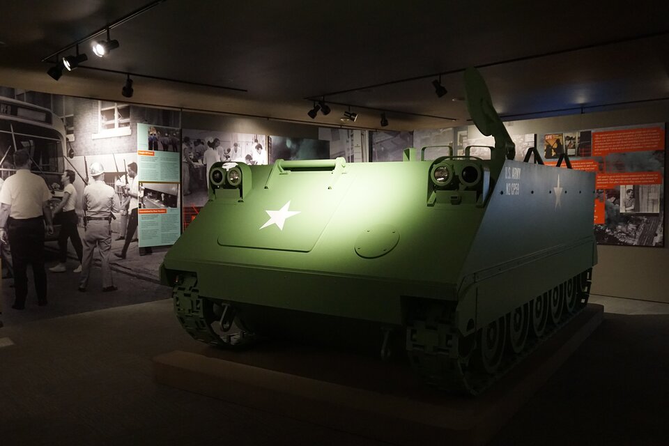 Na zdjęciu znajduje się wystawa w Muzeum Historycznym Detroit. Na środku ukazanego niedużego pomieszczenia stoi wóz opancerzony w kolorze zielonym, z białą gwiazdą na przedniej klapie. Z tyłu na ścianach znajdują się fotografie i tablice informacyjne.  