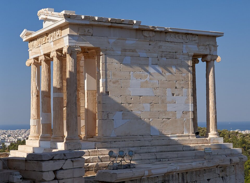 Zdjęcie przedstawia niewielką, zniszczoną starożytną świątynię. Prowadzą do niej niewielkie schody. Ukazany jest bok świątyni złożony z marmurowych bloków i jej front ozdobiony czterema kolumnami jońskimi podpierającymi zniszczony częściowo fronton. W oddali zabudowania miasta i niebo.