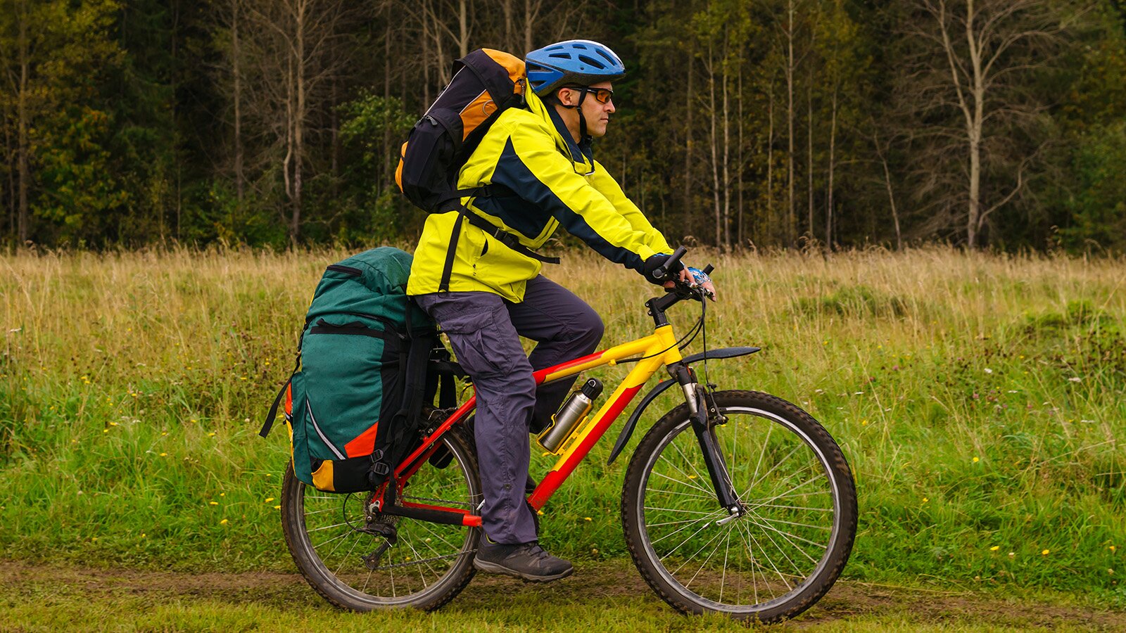 Zdjęcie przedstawia mężczyznę jadącego rowerem po drodze prowadzącej przez łąkę. Mężczyzna ma na plecach plecak, a nad tylnym kołem dużą wypchaną torbę.