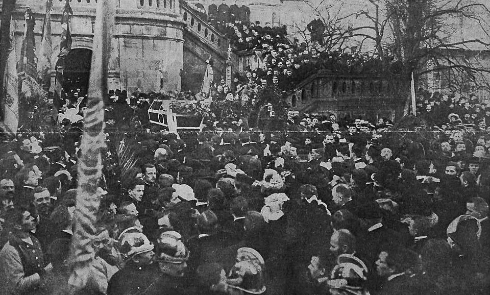 Czarno‑białe zdjęcie przedstawia tłum ludzi podczas pogrzebu. W centrum widać trumnę. Ludzie stoją na placu oraz na szerokich schodach w tle. Nad głowami żałobników widać uniesione sztandary.