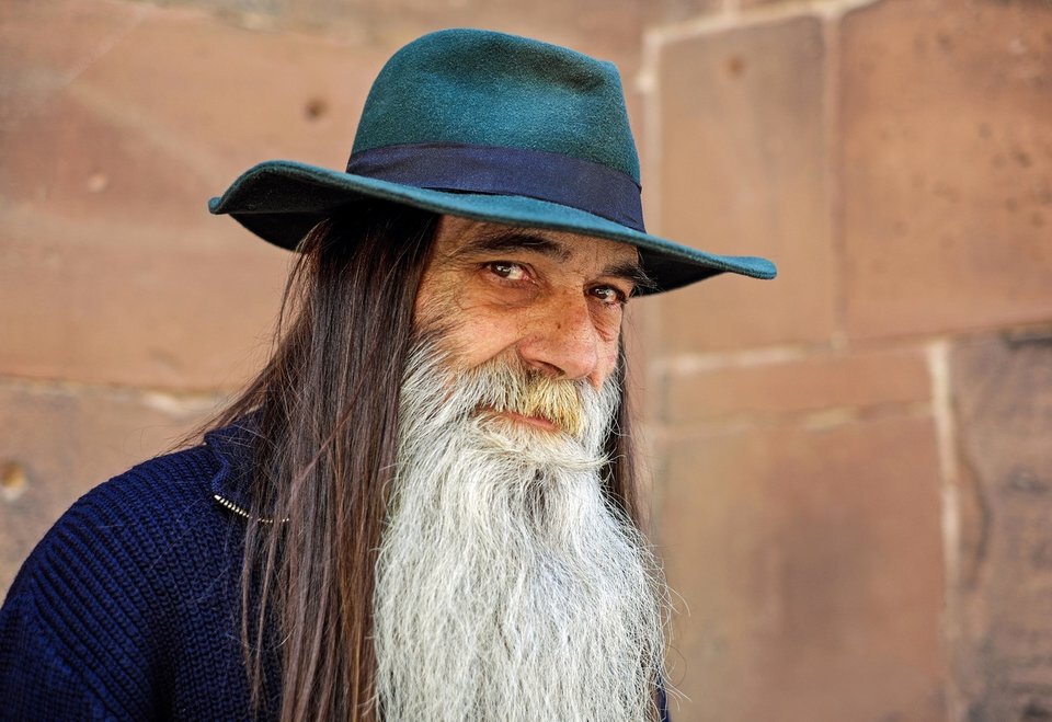 Fotografia kolorowa przedstawia starszego mężczyznę z długą siwą brodą, spod kapelusza wystają równie długie ciemne włosy. Mężczyzna spogląda w obiektyw kamery spokojnie, jakby dopiero powstrzymał uśmiech. W tle widoczna ściana z wielkich cegieł.