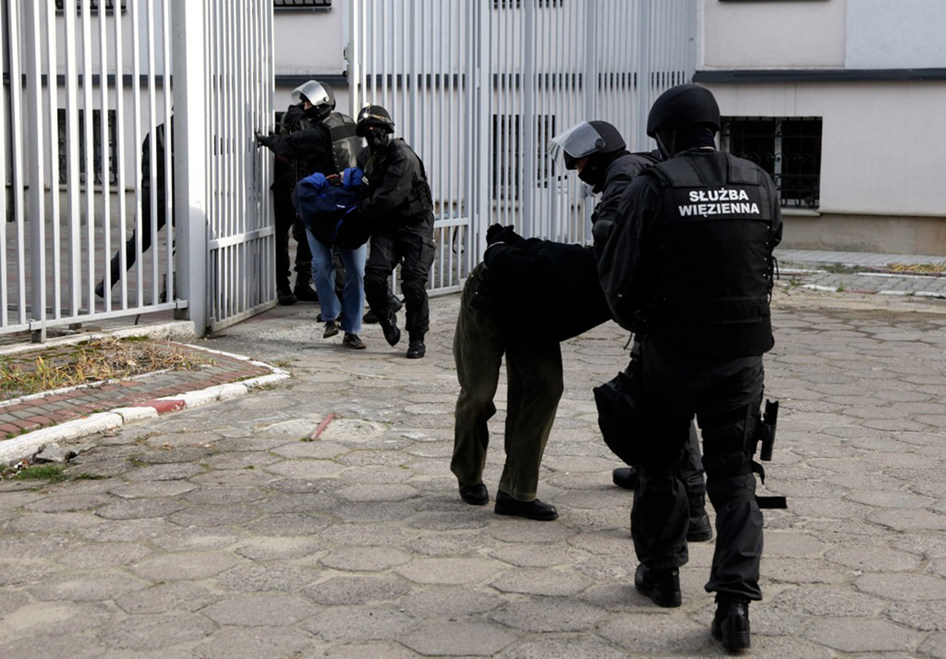  Kolorowe zdjęcie przed bramą więzienia. Wysokie metalowe ogrodzenie. Ogrodzenie składa się z równolegle ułożonych prętów. W części środkowej ogrodzenia lekko otwarta brama. Przed bramą funkcjonariusze służby więziennej. Na pierwszym planie dwaj funkcjonariusze tyłem do obserwatora zdjęcia. Ubrani w czarne mundury. Na głowie czarne kaski z przeźroczystą plastikową przyłbicą uniesioną do góry. Jeden z funkcjonariuszy prowadzi więźnia. Więzień jest pochylony do przodu, zgięty w pasie, dłonie na plecach skute kajdankami. W otwartej bramie dwaj następni funkcjonariusze prowadzący więźnia.
