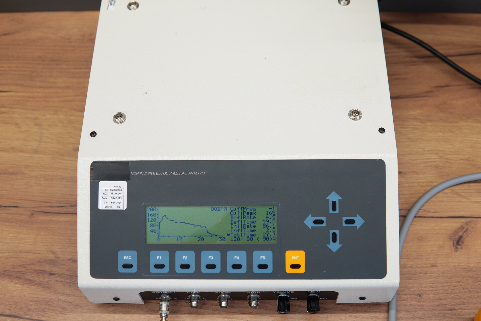 Zdjęcie przedstawia symulator nieinwazyjnego mierzenia ciśnienia tętniczego, w skrócie NIBP w perspektywie od góry. Na przedniej ścianie obudowy urządzenia znajduje się panel sterujący z przyciskami oraz ekranem, na którym widoczne są informacje o mierzonych wartościach pomiarowych oraz wykres. Pod panelem są gniazda do podłączenia urządzeń oraz dwa pokrętła. 