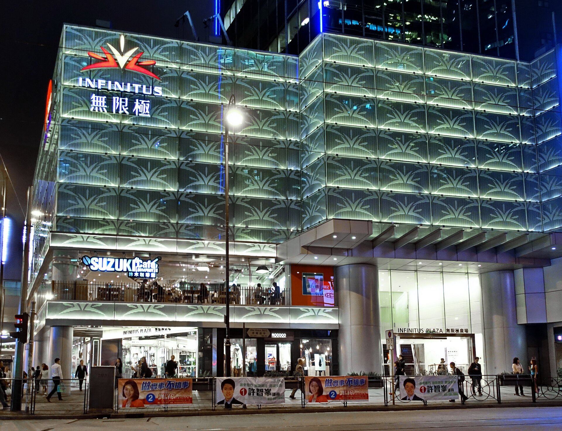 Fotografia przedstawia oświetlony budynek z czerwono‑białym logotypem przypominającym trzy ułożone obok siebie kwiaty – czerwony, biały oraz czerwony, z napisem „INFINITUS” tuż pod nim. Pod spodem widać litery chińskie. Większa część budynku jest w kolorze zielonym, z jaśniejszymi logotypami budynku umieszczonymi w sześciu rzędach. Parter oraz pierwsze piętro posiadają przezroczyste szyby, a pomiędzy nimi również widać zieloną ścianę z logotypami. Konstrukcja oparta jest na grubych filarach. Przed budynkiem, a także w środku, widocznych jest wiele osób. Na barierkach znajdujących się przed budynkiem oddzielających przejście od drogi znajdującej się przed barierkami umieszczone są plakaty reklamowe z podobiznami mężczyzny oraz kobiety.