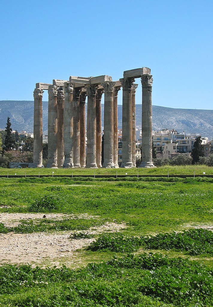 Zdjęcie przedstawia ruiny świątyni Zeusa Olimpijskiego w Atenach. W centralnej części zdjęcia widocznych jest trzynaście kolumn korynckich. Są one zwieńczone głowicami z bogato rzeźbionymi liśćmi akantu. Tło ruin stanowią ukazane w oddali współcześnie zabudowania Aten..