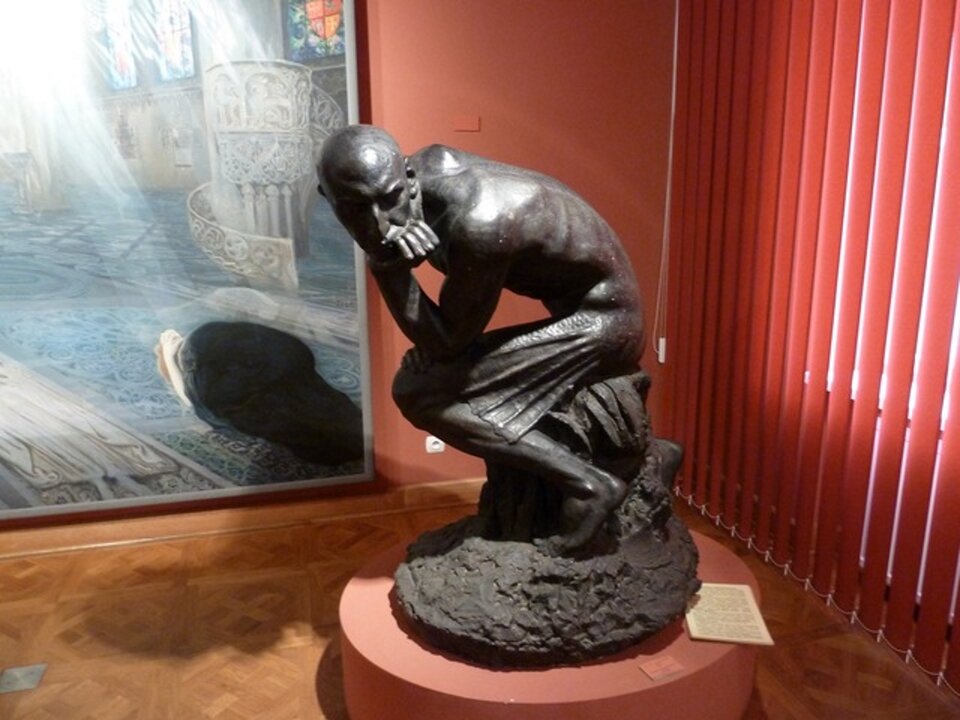 Ilustracja przedstawia rzeźbę pt. „Demon‑Wodnik” autorstwa Konstantyna Laszczki. Łysy mężczyzna siedzi i podpiera brodę o lewą rękę.