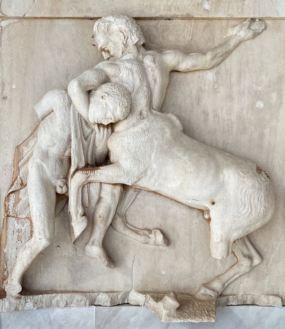 Ilustracja przedstawia Lapitę walczącego z centaurem, metopę południową I z Partenonu w Atenach. Ukazana jest dynamiczna scena walki wyrzeźbiona w marmurze: centaur w półobrocie unosi się na tylnych kończynach, przyciskając do siebie pochylonego wojownika, który odchyla się do tyłu w obronnym geście. Obie postacie są częściowo uszkodzone — brakuje głów i fragmentów kończyn — lecz zachowane partie ukazują silne napięcie mięśni i głębokie podcięcia charakterystyczne dla wysokiego reliefu. Tło stanowi gładka płyta metopy, a całość nosi ślady erozji.