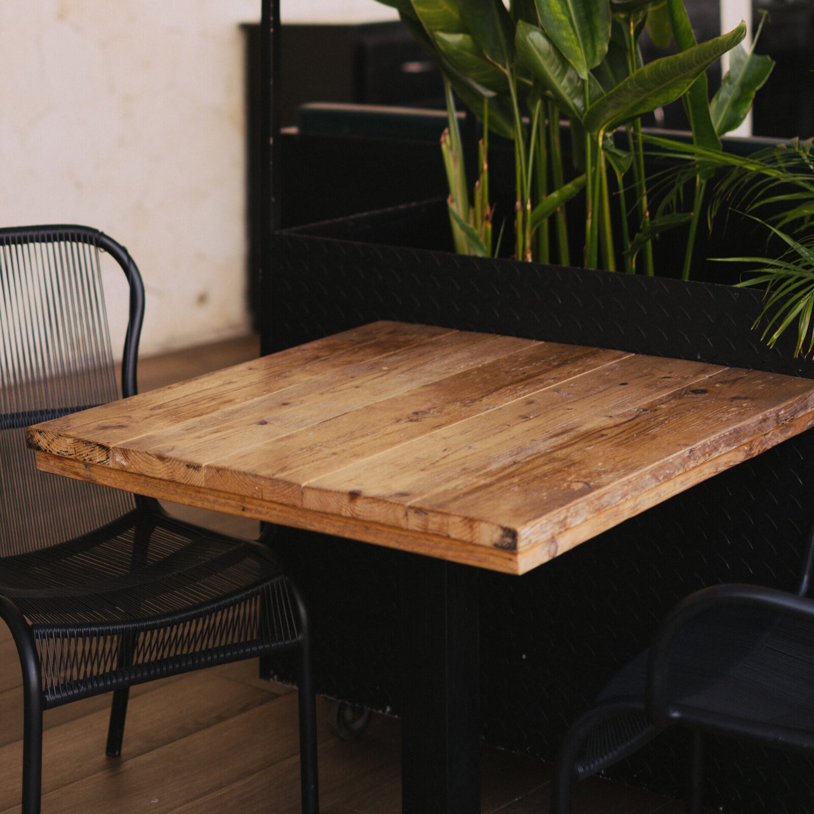 Ilustracja przedstawia mały kwadratowy stolik. Jest on drewniany, obok stoją czarne krzesełko i doniczka z kwiatami.