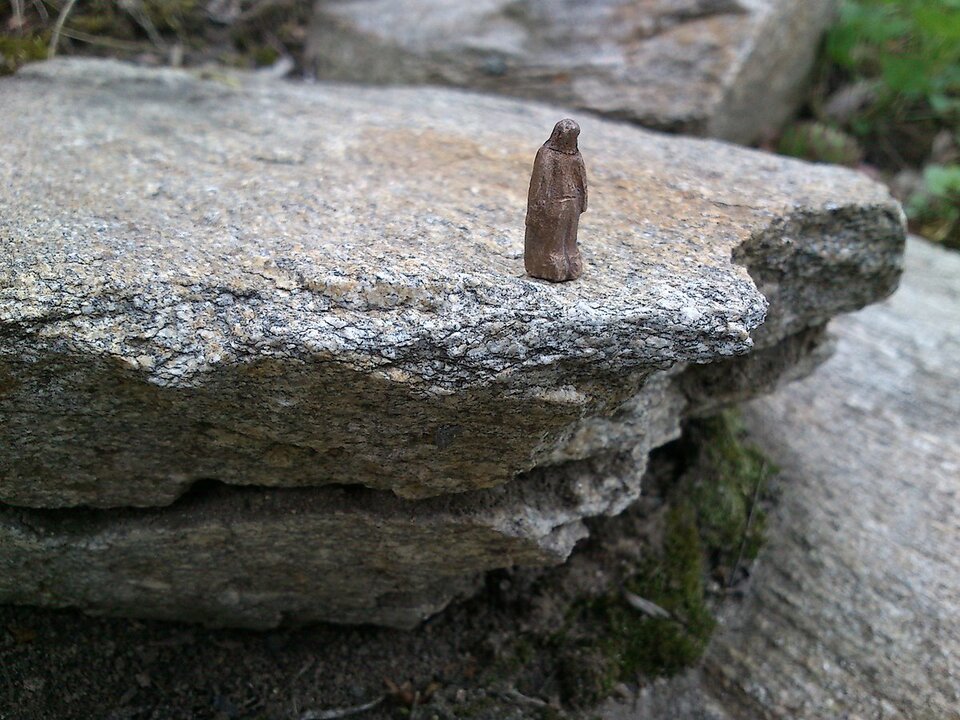 Ilustracja przedstawia niewielkich rozmiarów rzeźbę św. Barbary, stojącą na skraju płaskiej półki skalnej. Skała wydaje się ogromna w stosunku do miniaturowej statuetki. 