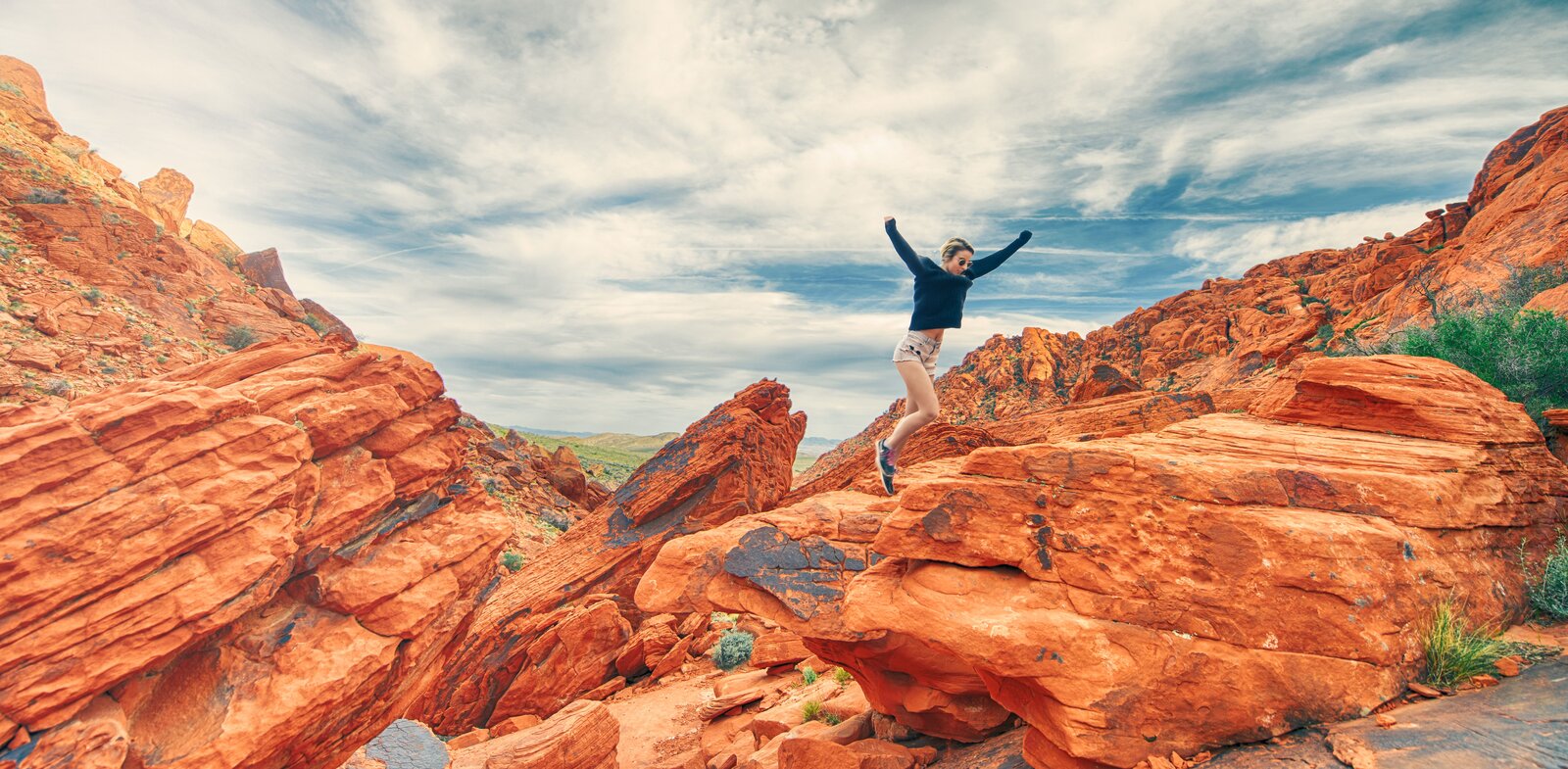 Fotografia przedstawia pomarańczowe skały, wśród których skacze dziewczyna ubrana w krótkie spodenki i sweter. Pomiędzy niektórymi skałami wyrastają zielone trawy. Na niebie widoczne są białe obłoki.