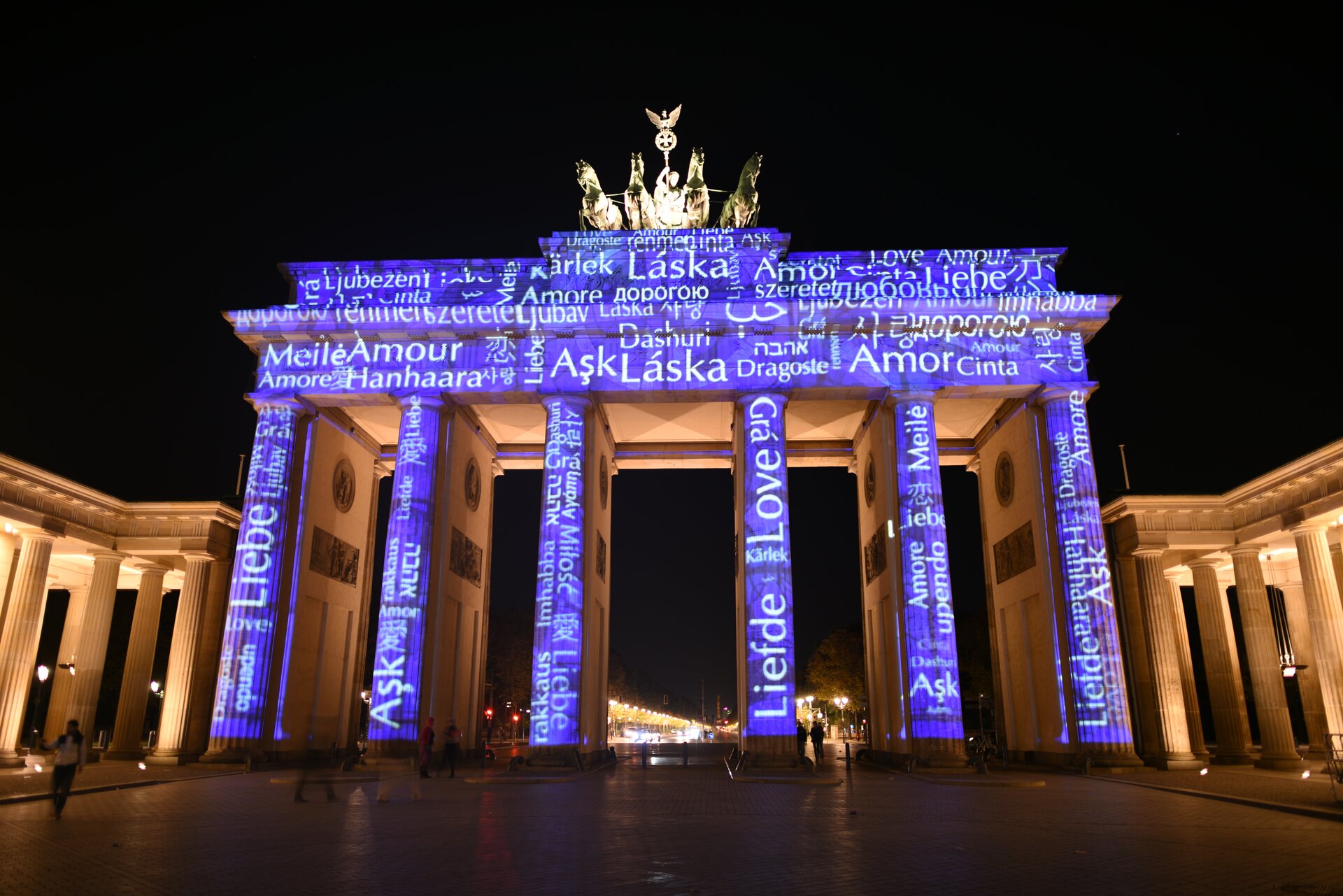 Na zdjęciu znajduje się beżowa budowla - Brama Brandenburska, która konstrukcyjnie opiera się na kolumnadzie złożonej z sześciu wysokich kolumn. Na jej zwieńczeniu znajduje się posąg z 4 końmi. Fioletowe tło oraz białe napisy wyświetlane są na kolumnach i ścianach budynku. Na placu, na którym stoi jest kilka postaci. Zdjęcie wykonano w nocy. 