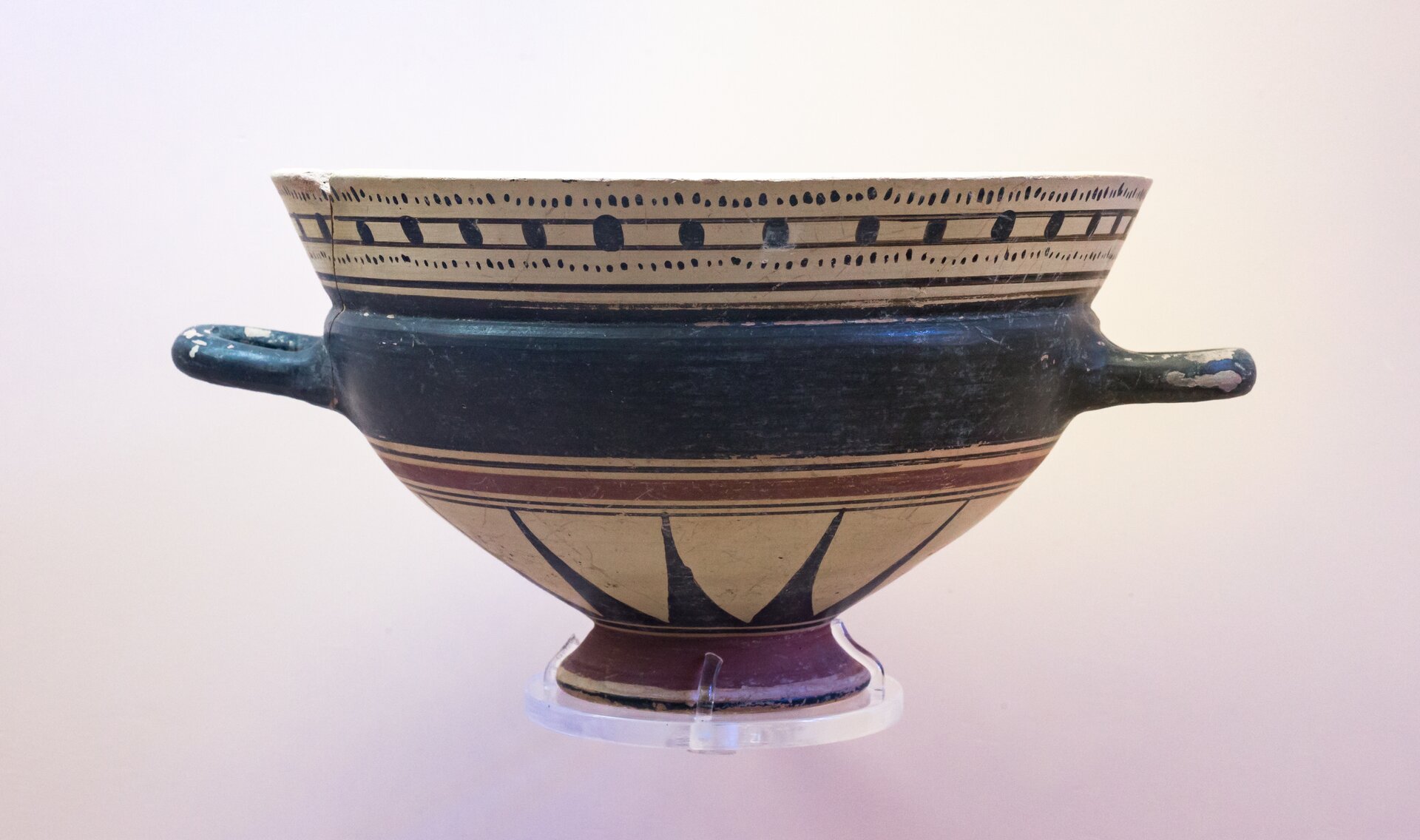 Fotografia autorstwa ArchaiOptix przedstawia starożytny kyliks - naczynie kształtem przypominającą dzisiejszą wazę obiadową. Jest to szerokie naczynie zwężające się u podstawy. Z obu stron, mniej więcej w połowie wysokości naczynia znajdują się uszy służące do jego przenoszenia. Waza jest trójkolorowa, szyjka zdobiona jest czarnymi wzorami geometrycznymi na beżowym tle, u dołu namalowane są trójkąty również na beżowym tle. Środkowa część wazy ma jednolity ciemny kolor.