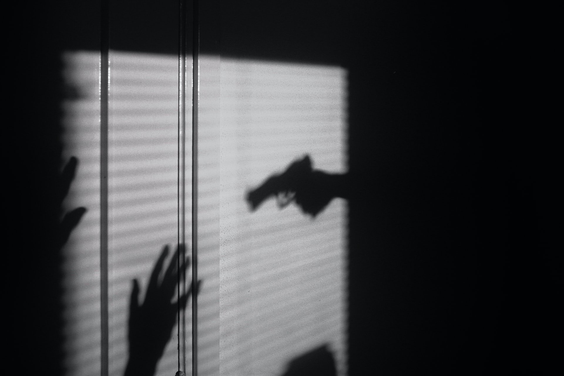 Zdjęcie przedstawia ciemne pomieszczenie, do którego wpada światło. Na jego jasnym tle znajdują się uniesione do góry dłonie i skierowana w jej kierunku dłoń z pistoletem. 