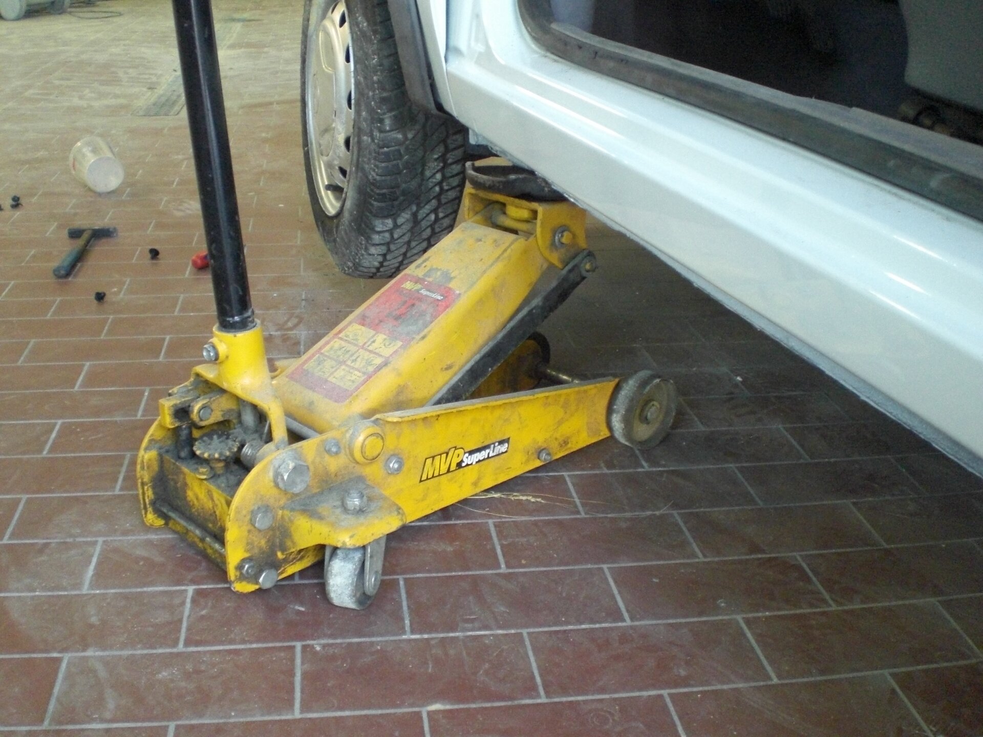 Na ekranie widoczne wnętrze warsztatu samochodowego. Na podłodze urządzenie składające się z ramy na kółkach z uniesionym elementem środkowym, który wsunięty jest pod samochód i służy do jego unoszenia.