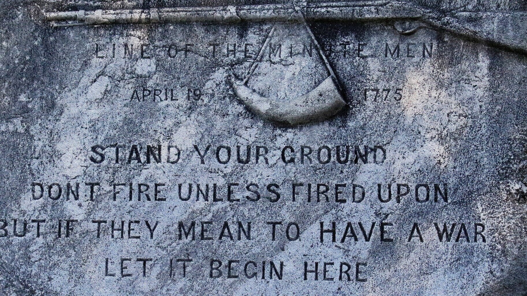 Fotografia barwna. Kamienna, ciemnoszara tablica. U góry płaskorzeźba przedstawiająca muszkiet. Na jego długiej lufie wisi przymocowany do sznurka róg na proch strzelniczy.  Poniżej wyryty tekst: LINE OF THE MINUTE MEN APRIL 19, 1775 STAND YOUR GROUND DON'T FIRE UNLESS FIRED UPON BUT IF THEY MEAN TO HAVE A WAR LET IT BEGIN HERE