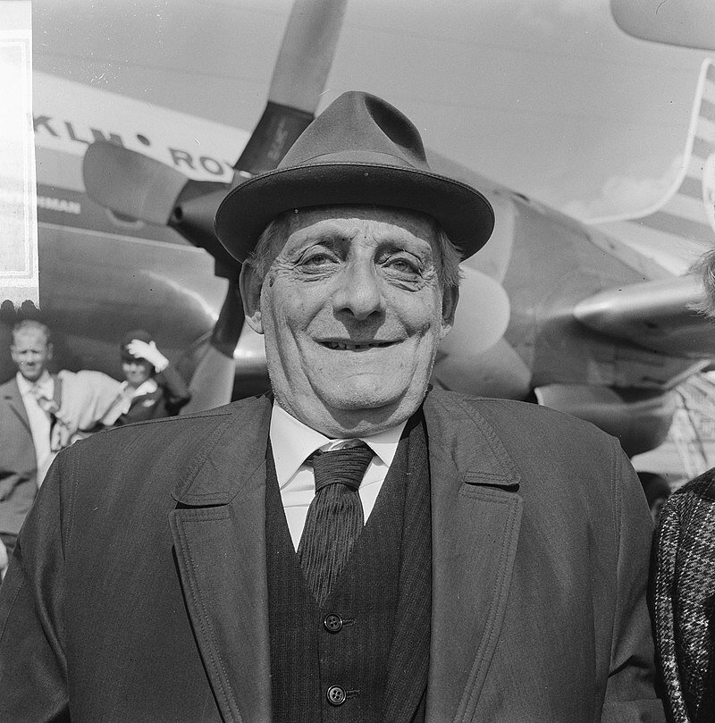 Zdjęcie przedstawia starszego mężczyznę w kapeluszu. Mężczyzna się uśmiecha. Ubrany jest w płaszcz, marynarkę, kamizelkę, koszulę i krawat. W tle samolot oraz mężczyzna i kobieta.